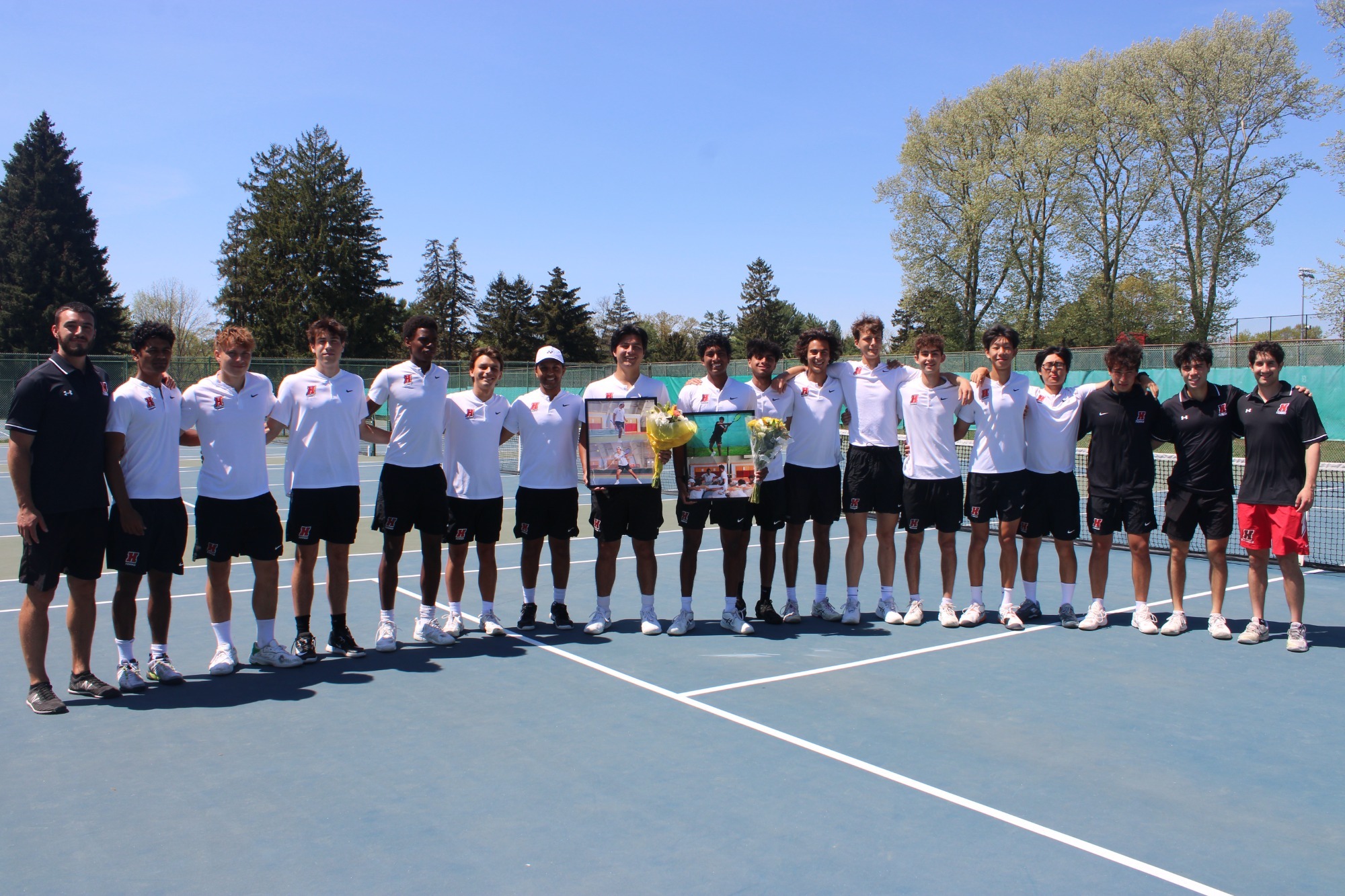  The Haverford men’s tennis team poses on court for Senior Day, honoring Stefan Johnson and Niki Lavu, who stand center holding commemorative photo frames and bouquets under a clear blue sky.