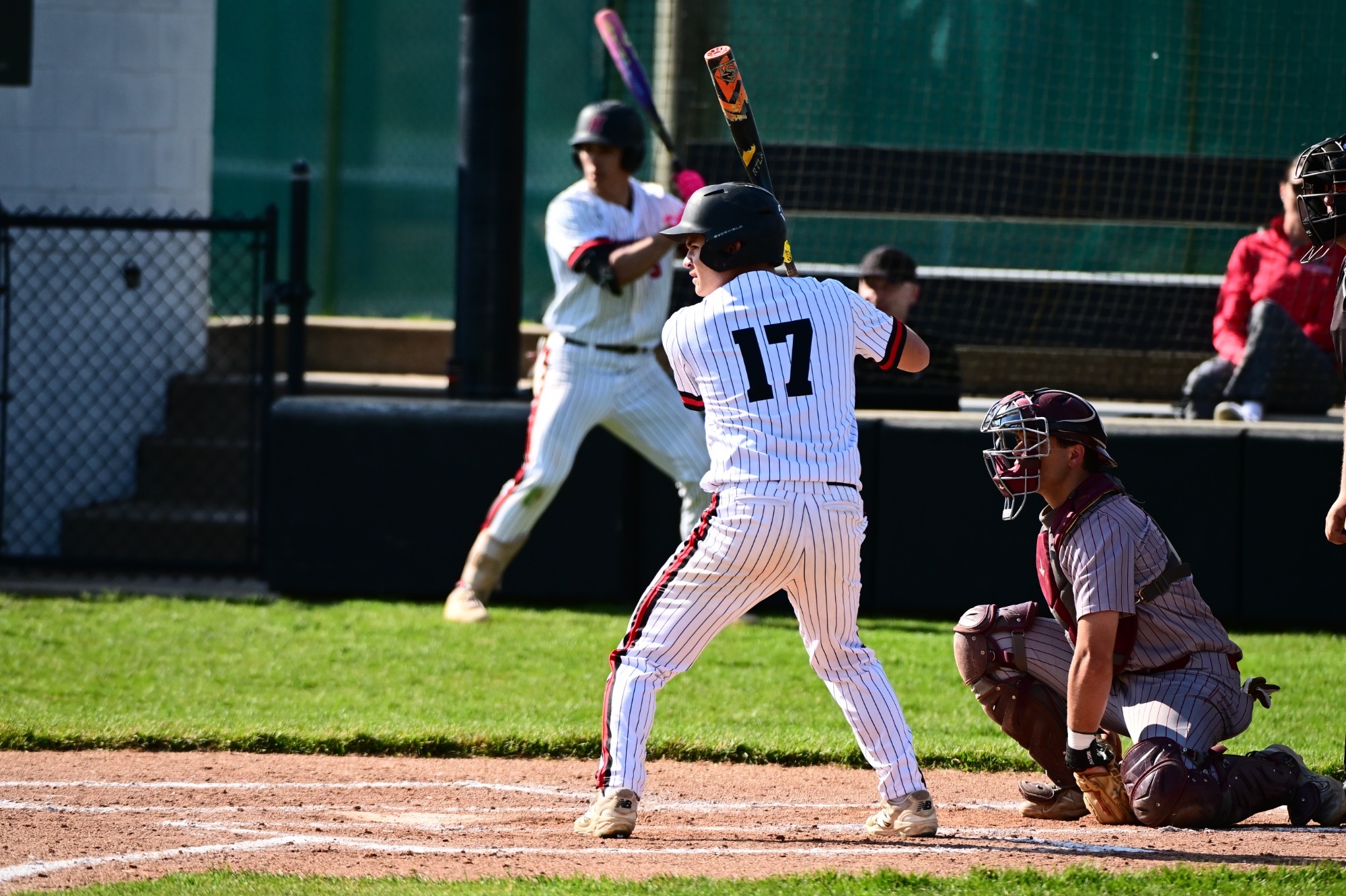 Sammy Angel wearing a white pinstriped jersey with the number 17 and a black helmet stands in a batting stance at home plate. He is holding a black and orange bat over his shoulder, focused on the pitcher. Behind him, a catcher in maroon gear is crouched in position, and another player is visible in the background on deck. The scene is captured on a sunny day at a baseball field with green grass and a dirt batter's box.