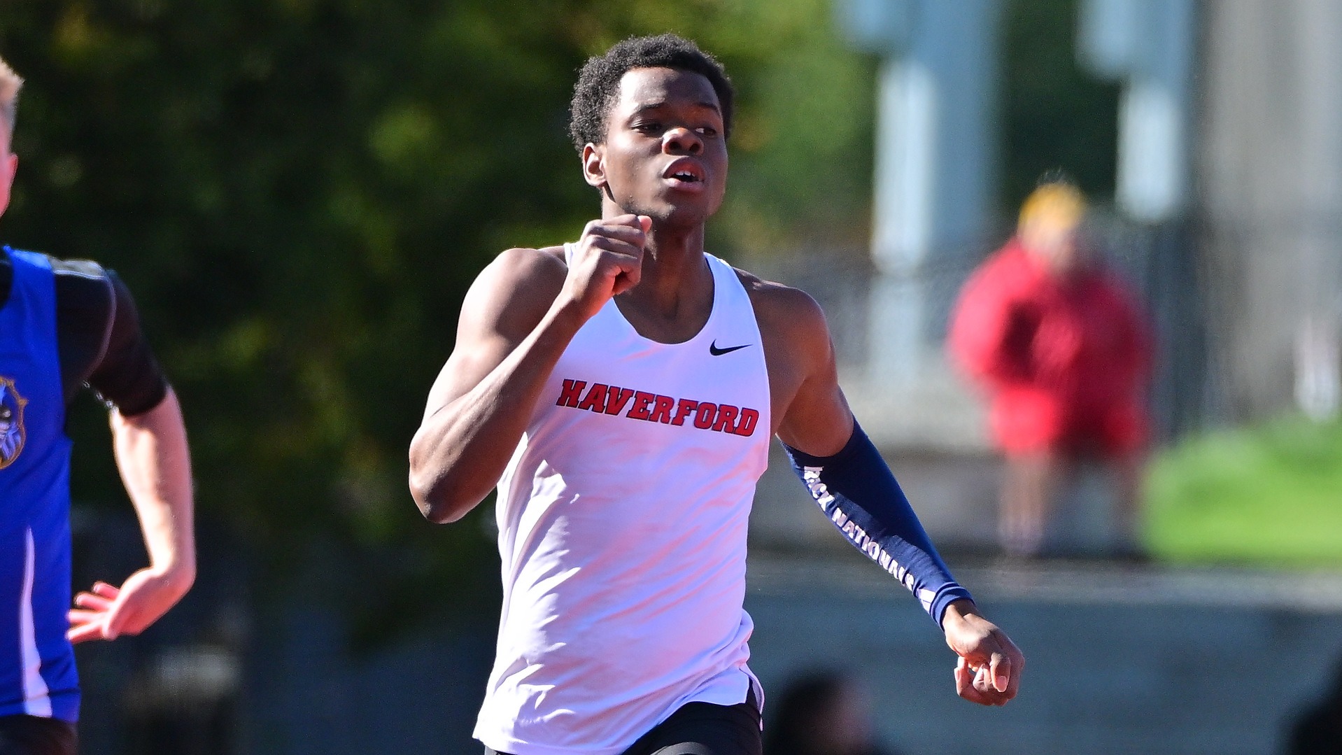 Josue sprints forward in a white Haverford College tank top with red lettering. He wears a navy blue compression sleeve on his left arm and maintains a focused expression during a race on a sunlit outdoor track.