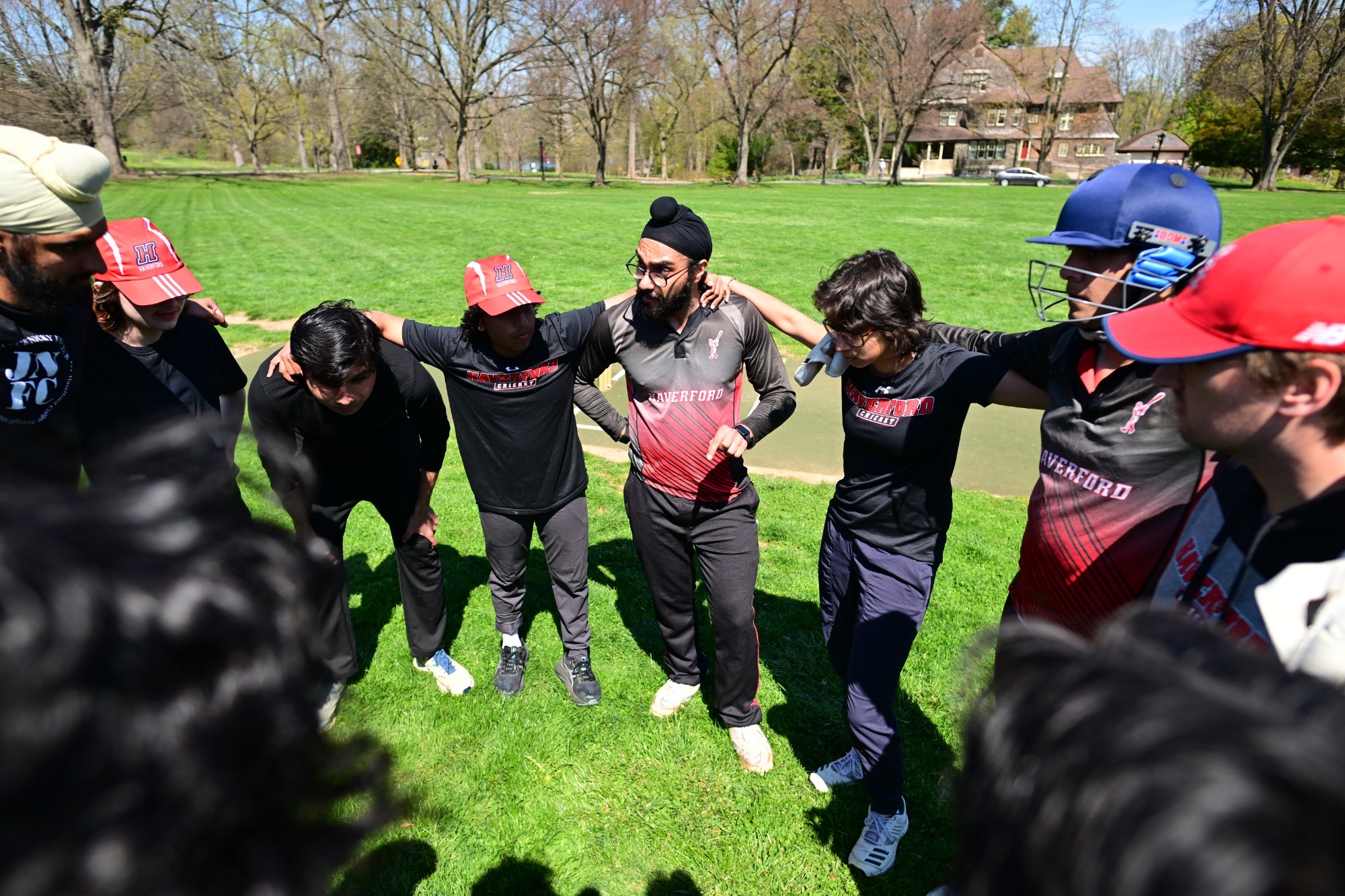 A huddle of the Haverford College cricket team on a sunny day. The players are gathered in a tight circle on a bright green field, with some wearing red and black Haverford-branded athletic gear and caps. At the center of the huddle, a player wearing a black turban and glasses appears to be addressing the group. In the background, the campus is visible with lush trees and a large stone building under a clear blue sky.