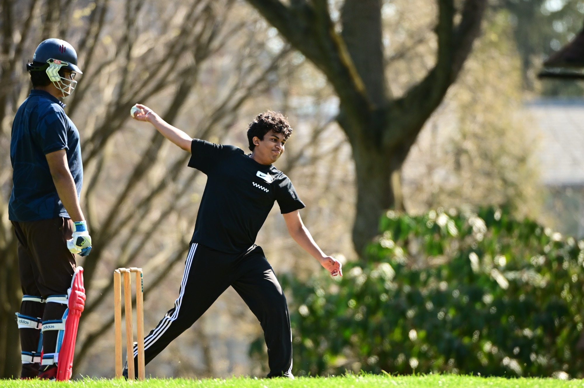 Anish More, in a black shirt and athletic pants, delivers a cricket ball during a match on a sunlit field.