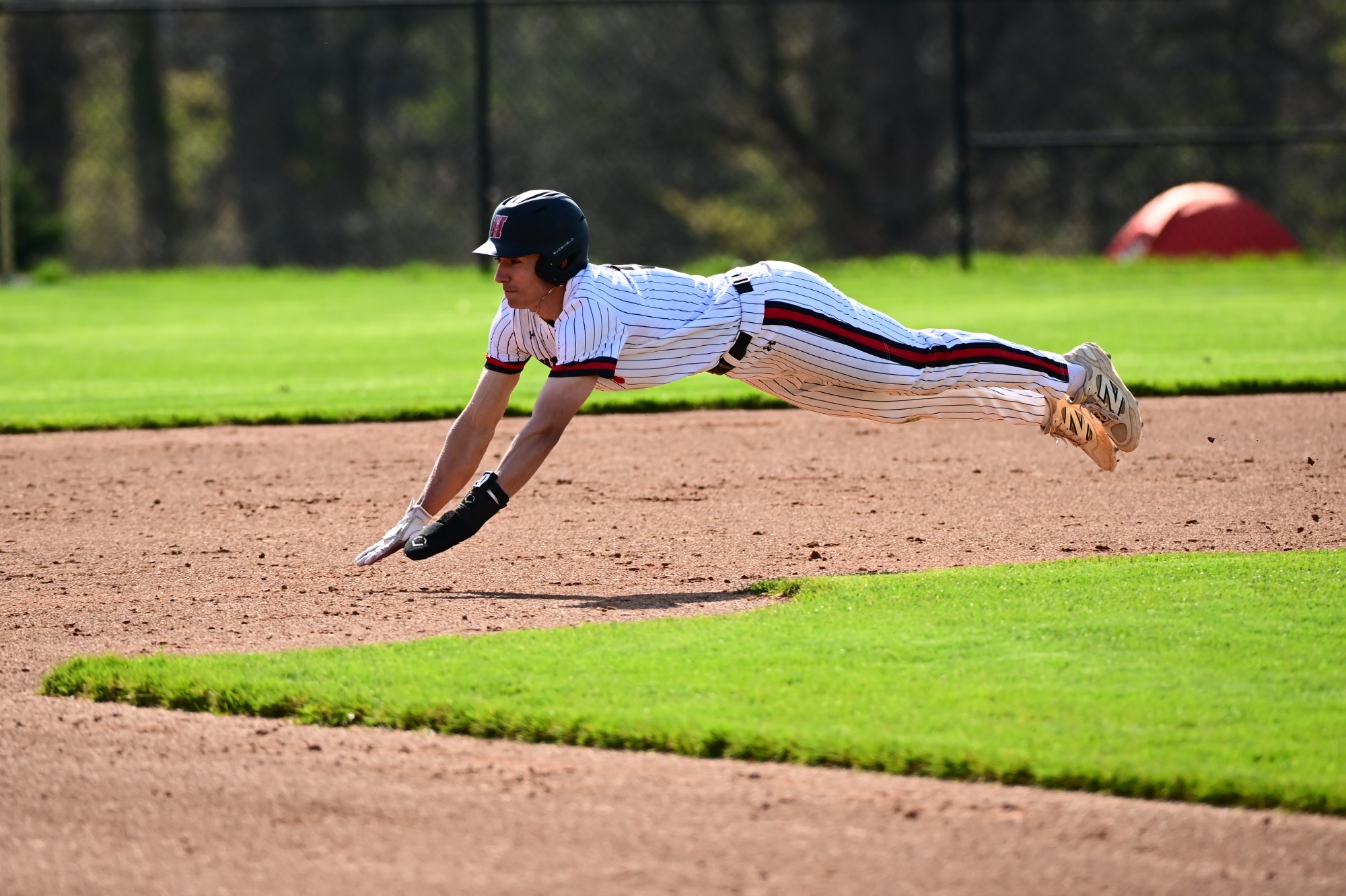 Baseball player Mauro DeCillis, in a pinstripe uniform, fully airborne in a horizontal dive across the dirt toward a base during a sunny daytime game.