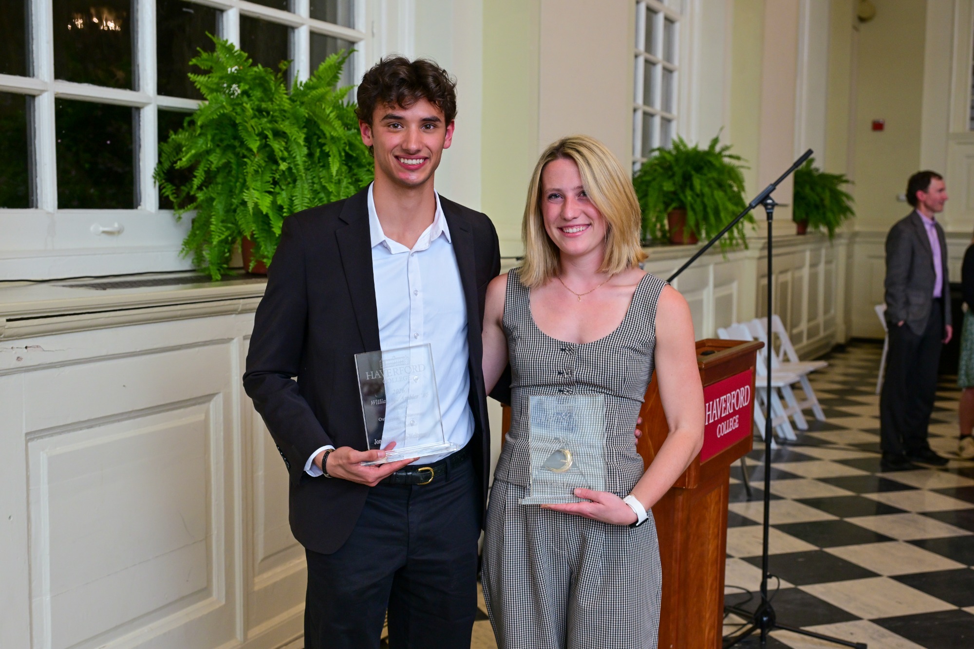 Carly Priest and Jonah Hoffmann-Weitsman stand side by side at an indoor awards ceremony, smiling and holding their Ambler Scholar-Athlete plaques. Priest wears a sleeveless patterned dress, while Hoffmann-Weitsman is dressed in a dark suit and light shirt. They pose in front of tall windows with green plants, with a podium and microphone visible nearby.