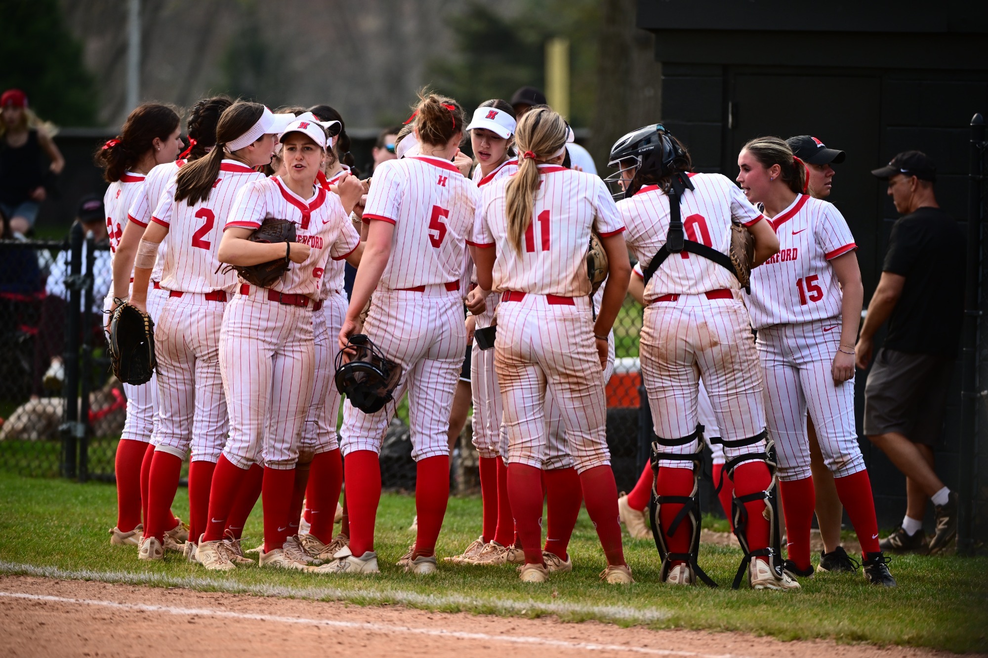 Haverford players in white pinstriped uniforms and red socks gather in a tight huddle on the field, dirt-stained and focused, while coaches and spectators stand in the blurred background.
