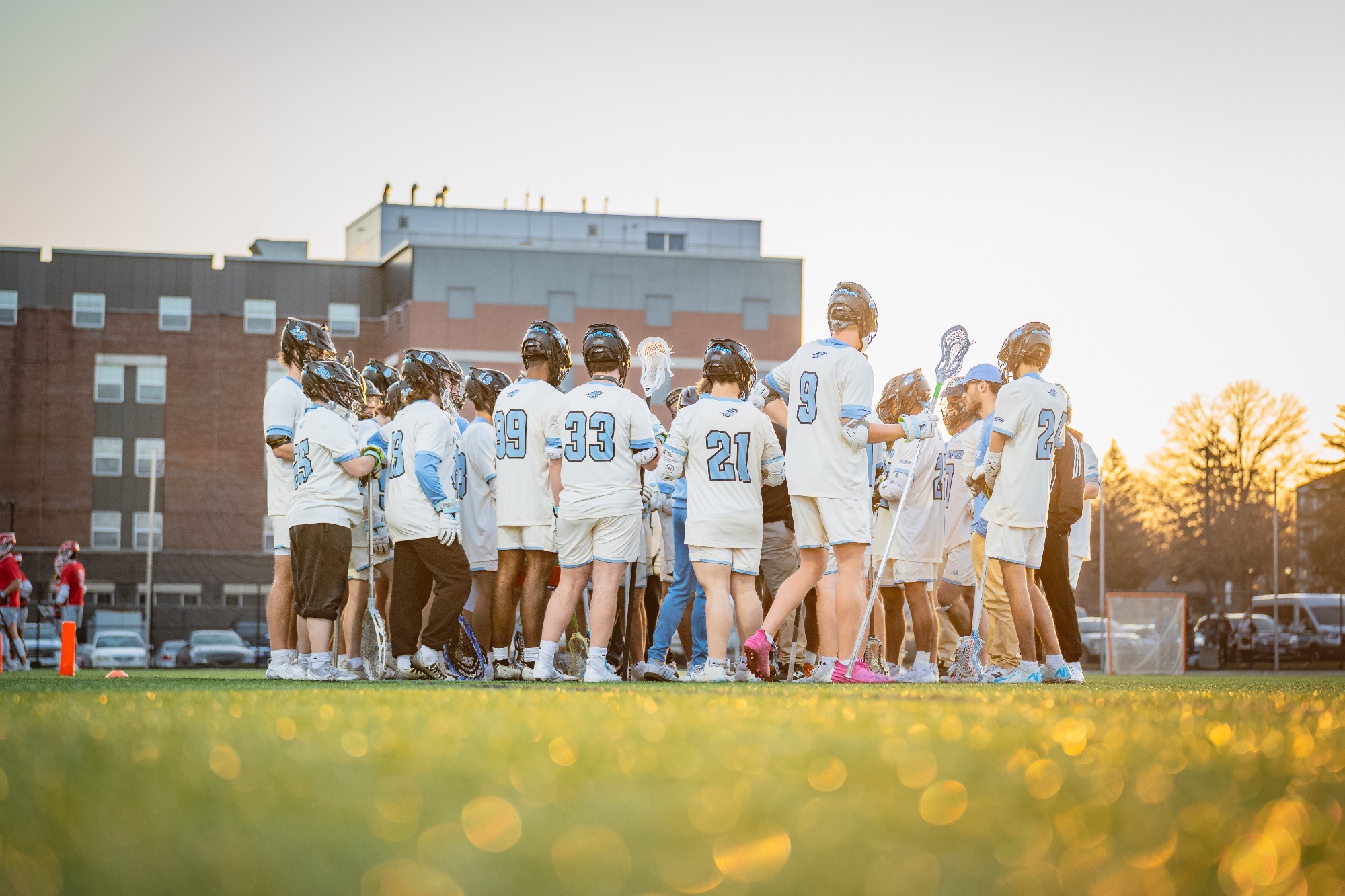 The 2025 HFU Men's Lacrosse Team huddles on the field, sun shining in the background