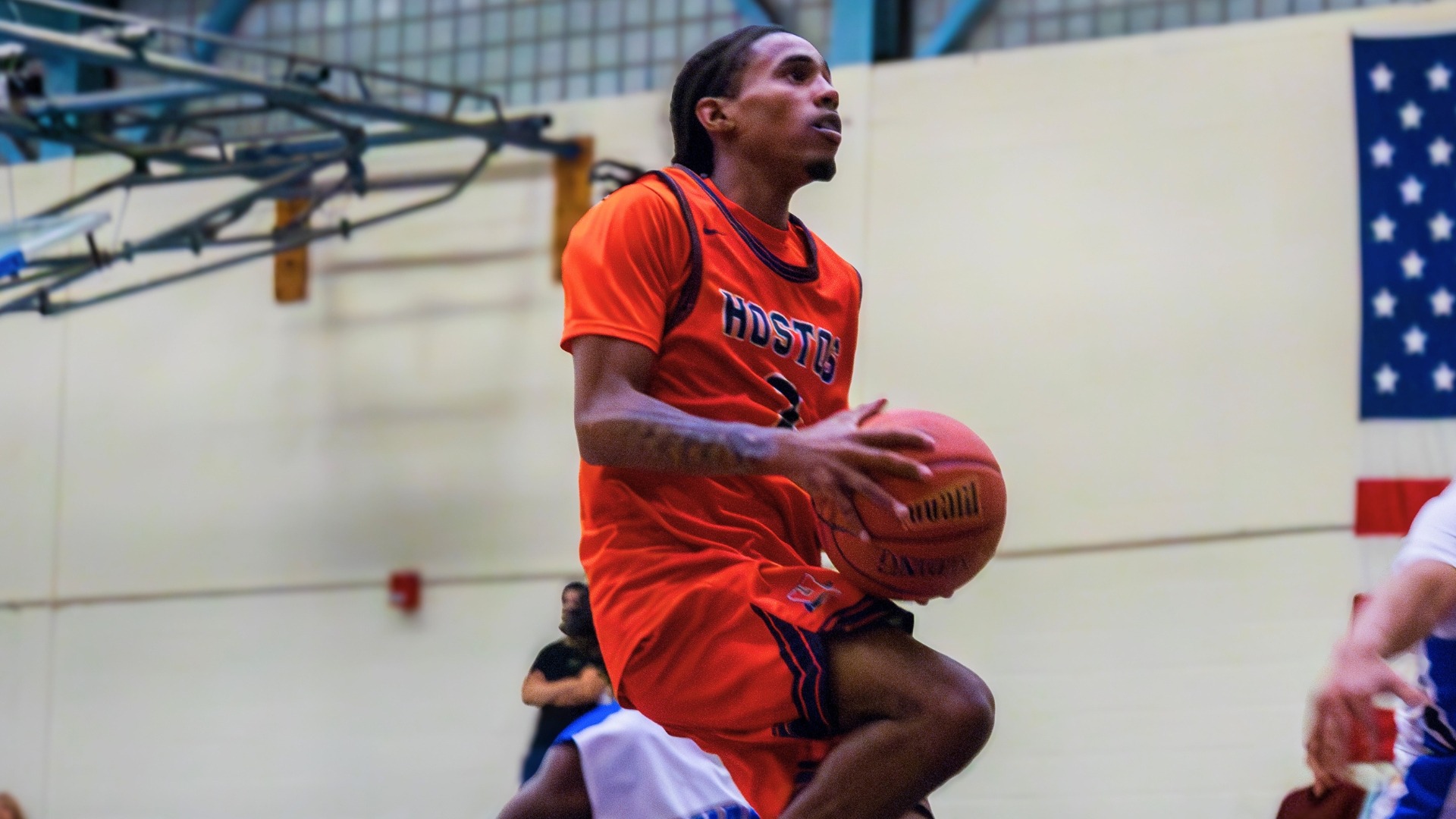 Hostos men’s basketball player in an orange uniform drives toward the basket while holding the ball, with a defender nearby and an American flag visible in the gym background.