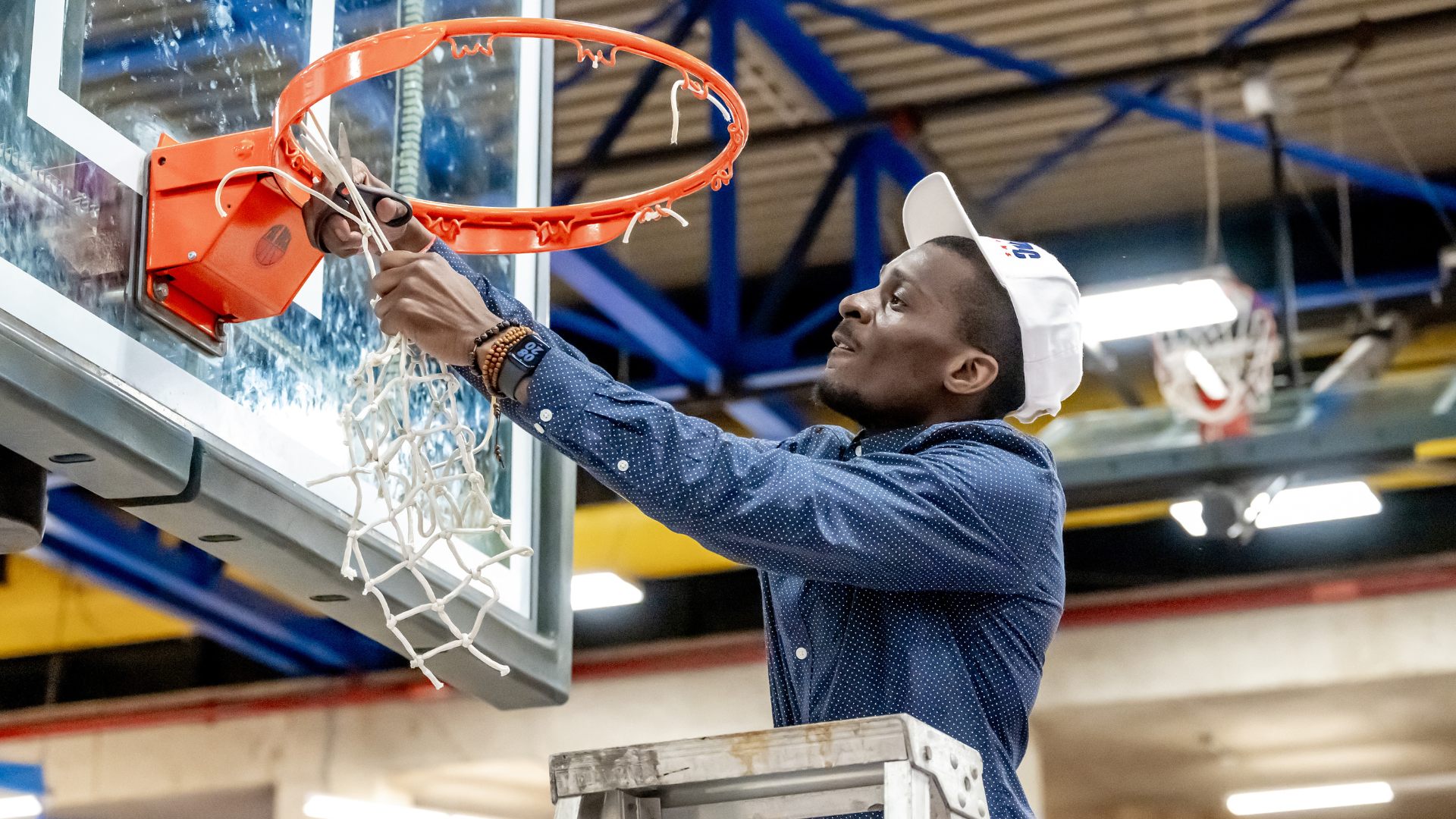 Coach Dwight Shaw cuts down the net while standing on a ladder following a championship victory, wearing a blue button-down shirt and white championship hat inside a gymnasium.