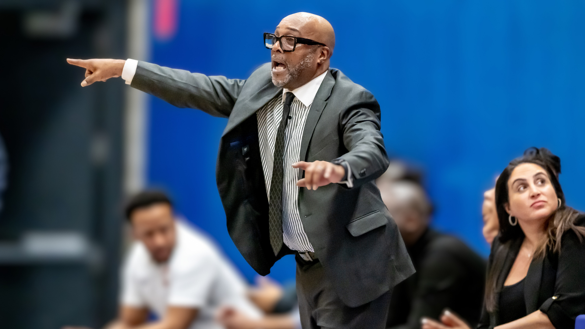 A basketball coach in a dark suit points across the court while calling instructions from the sideline, with staff members seated behind him during a game.