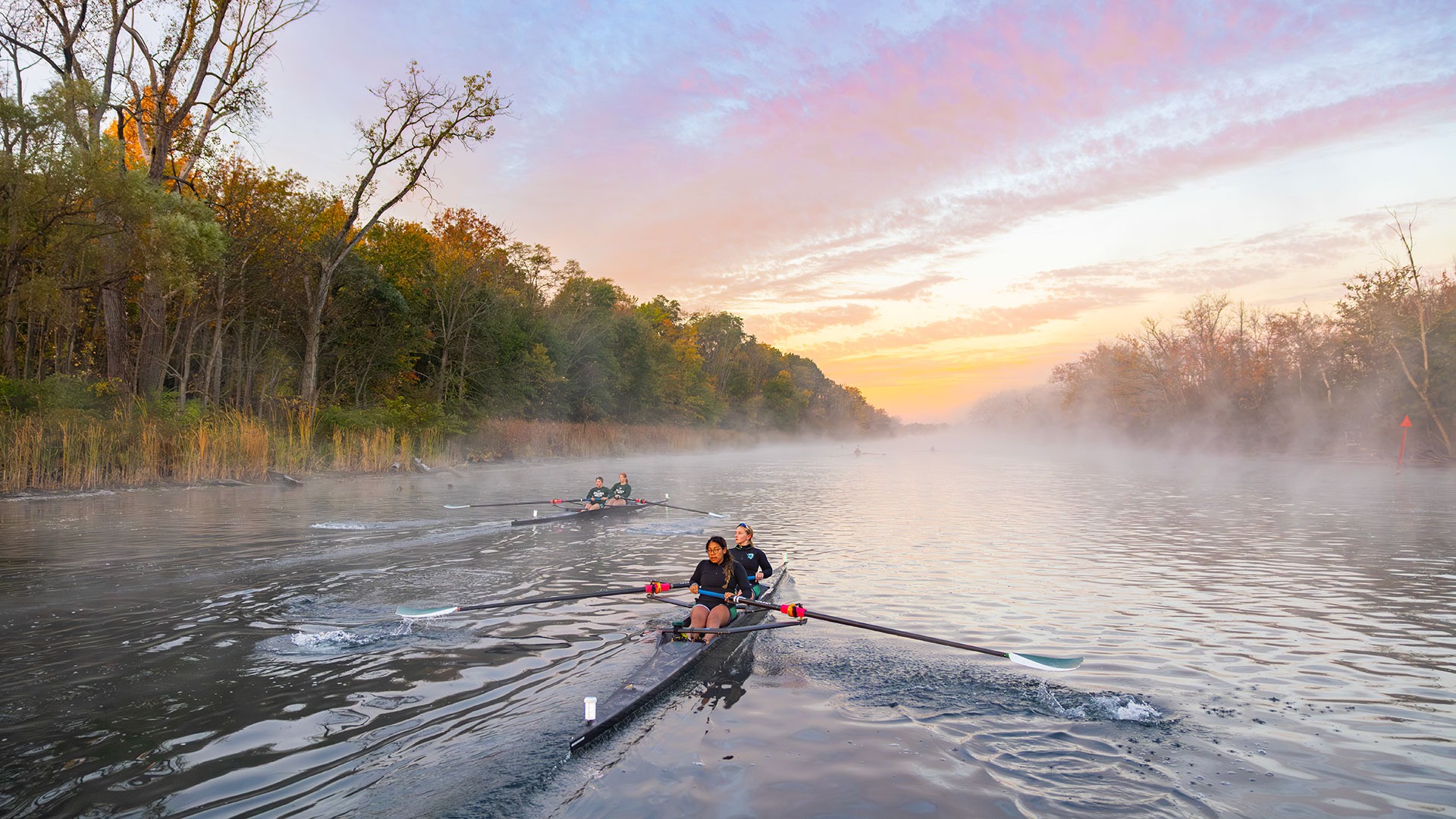 Two members of the William Smith rowing team sit on a boat. They are rowing into a misty morning sunrise on the Canal