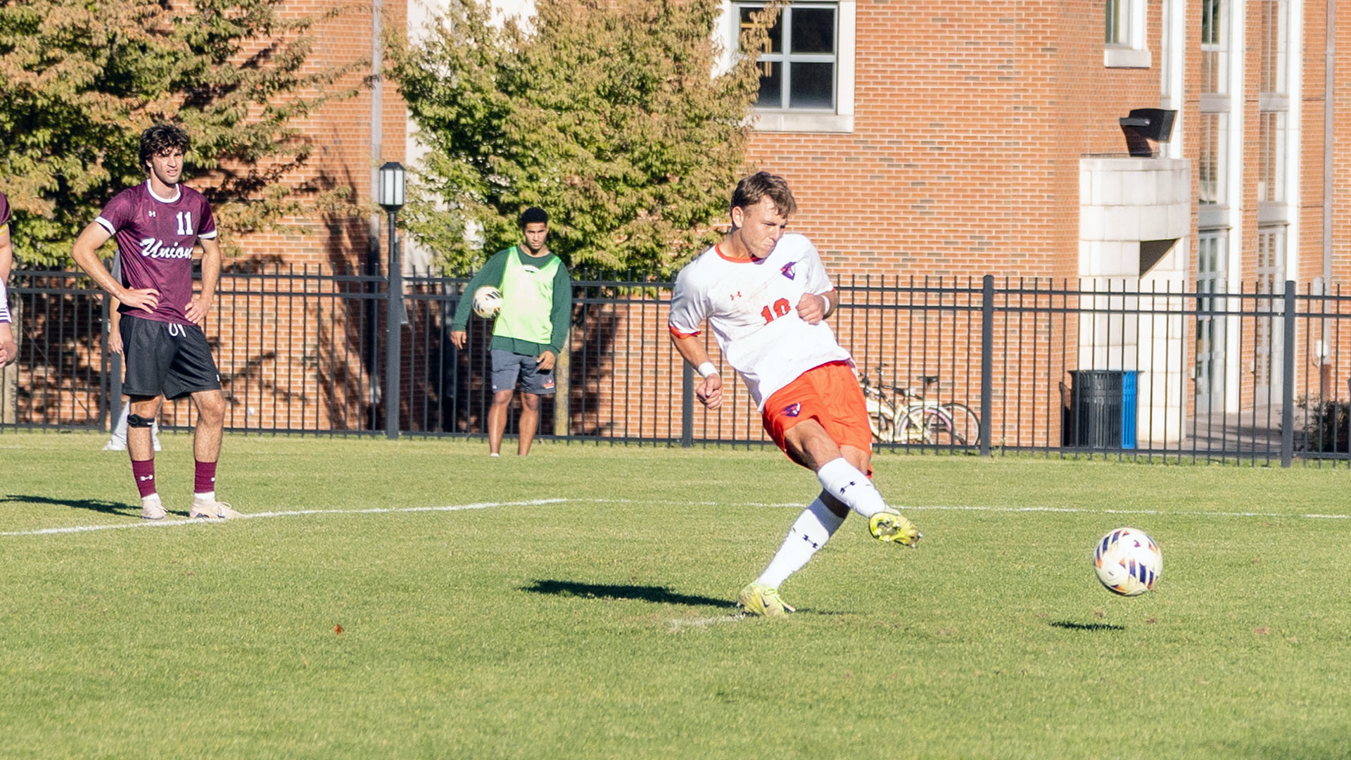 A male soccer player in a white jersey and orange shorts follows through on a penalty kick with an opposing player in a maroon jersey and black shorts standing in the background with hands on hips.