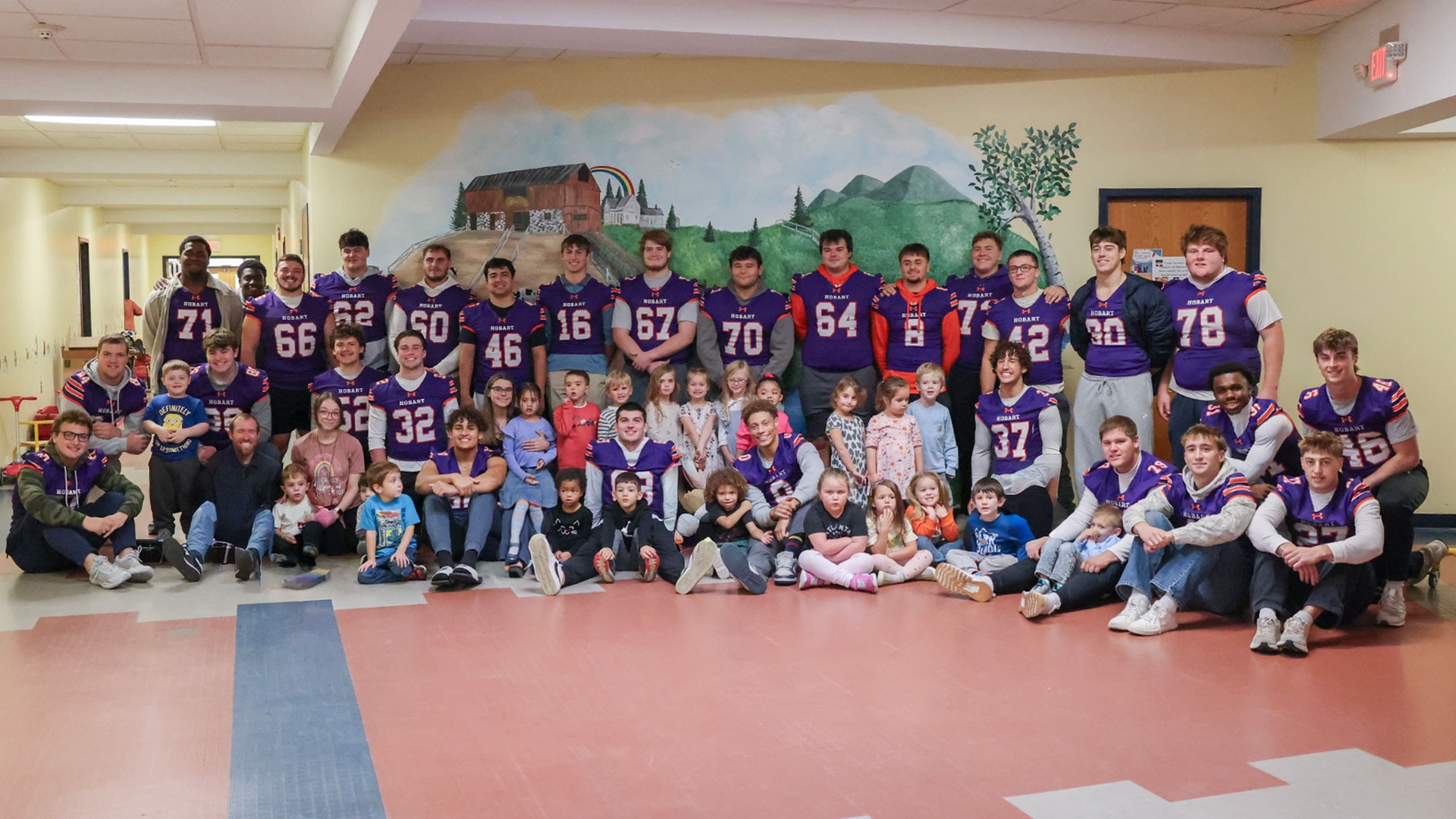 A large group of Hobart football players wearing their purple jerseys and students and staff from Happiness House pose for a picture in the school's hallway.