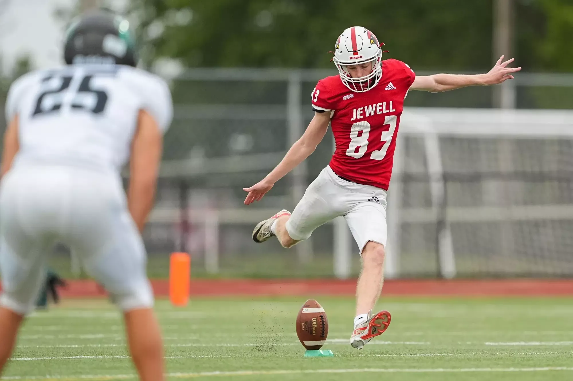 Football player in a red Jewell jersey and helmet kicks a football during a game, with an opposing player in a white uniform blurred in the foreground. The ball is positioned on a kicking tee on the field