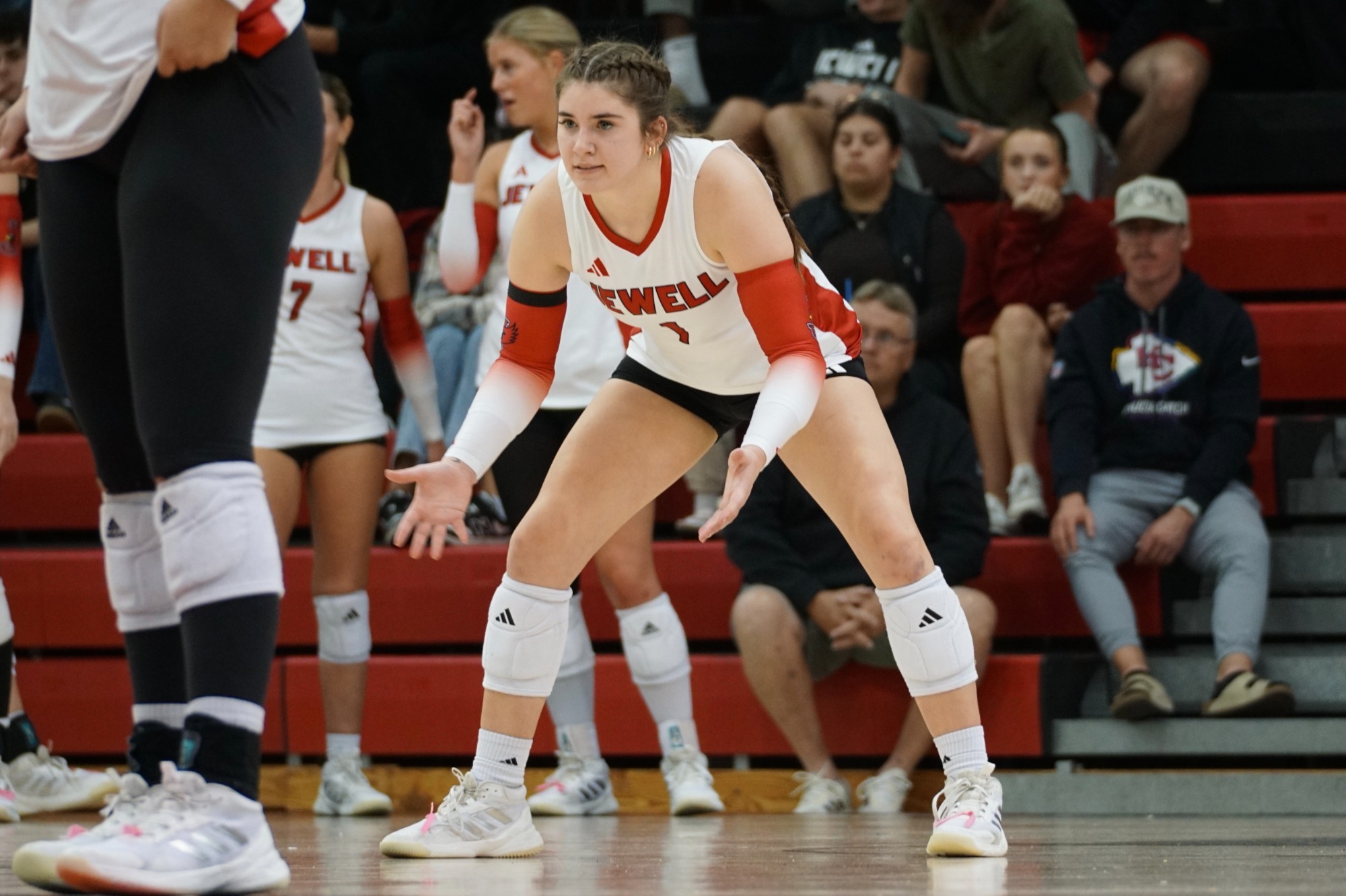 Ashlynn Rowland stands in a defensive stance with her arms out in front, wearing white jersey and kneepads. Fans are visible in the background.