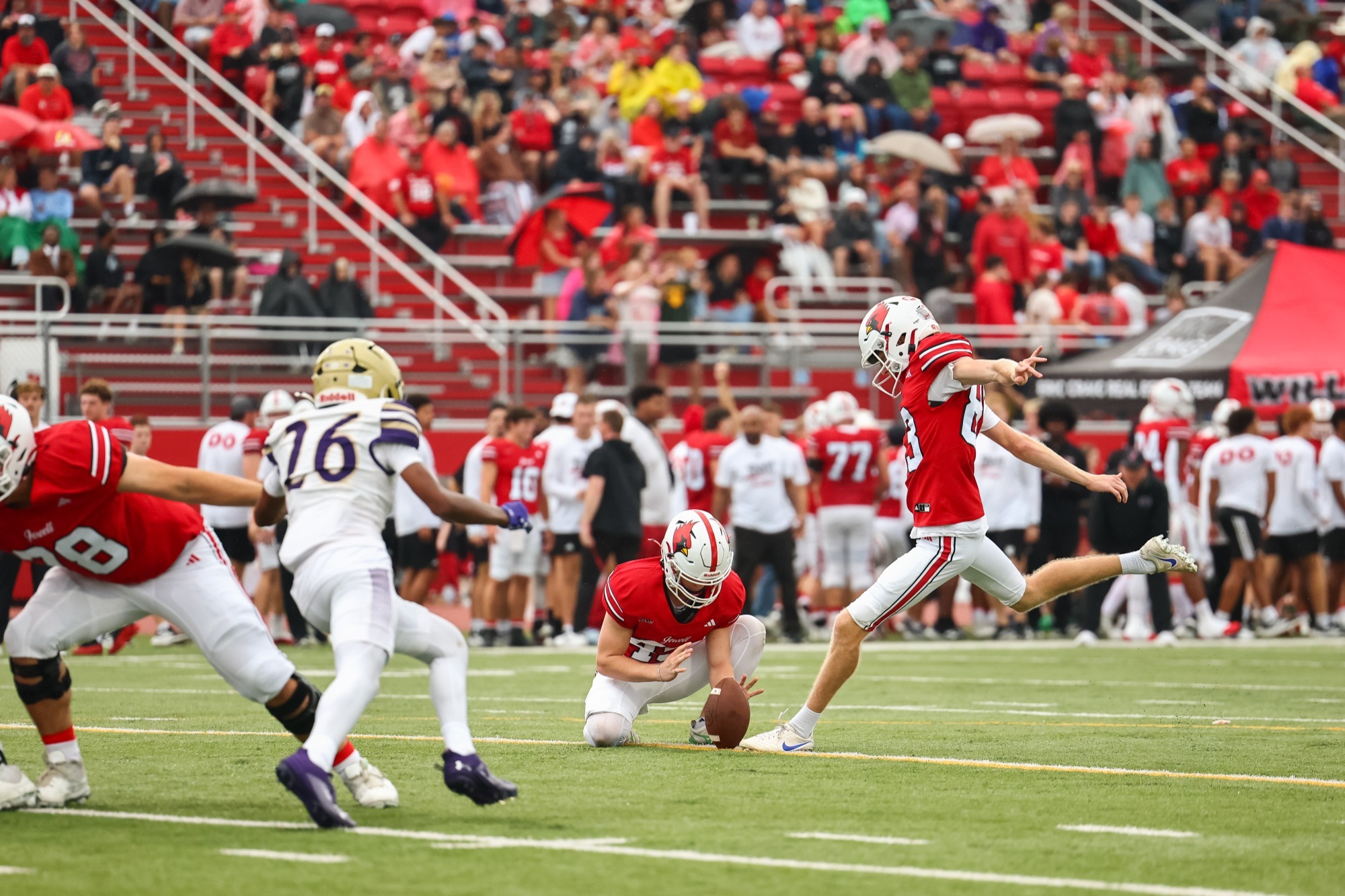 Bleu Renfrow kicks a field goal, with Frankie Kapetanovich holding. A McKendree player is running toward them in the foreground with the Jewell sideline and the stadium full of fans blurred in the background 