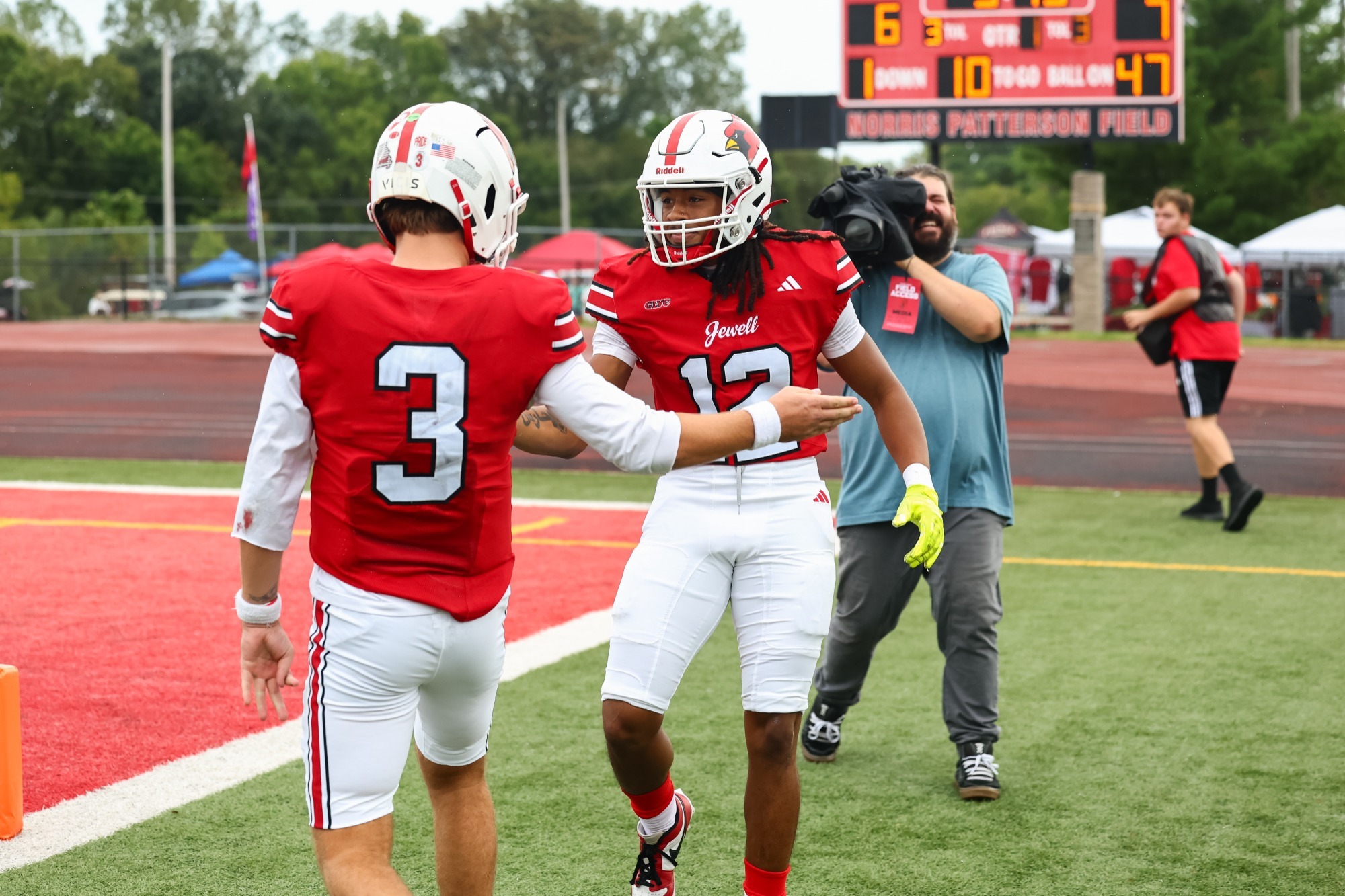 Xavier Winston high fives Charlie Loose on the sideline after scoring a touchdown. Both players are wearing red jerseys, white helmets, and white pants. A man with a news camera stands behind them with the scoreboard slightly blurred in the background.