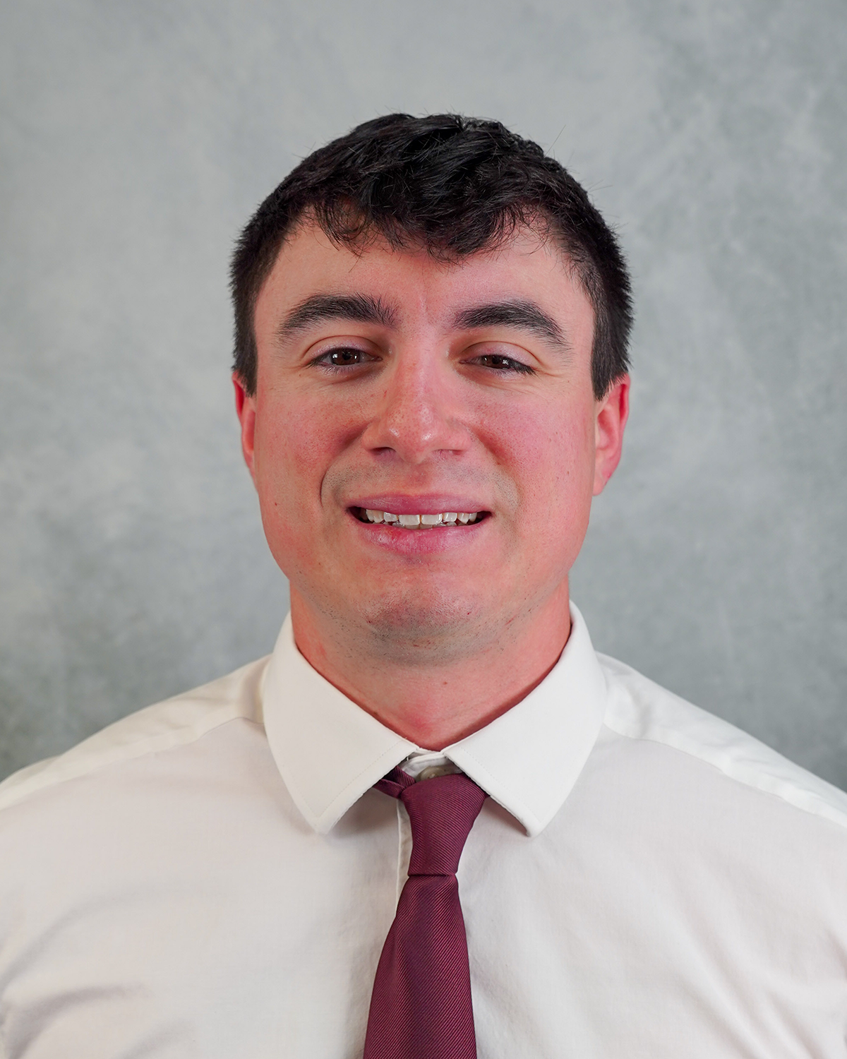 Andrew Briones poses for a headshot wearing a white shirt and red tie against a gray background