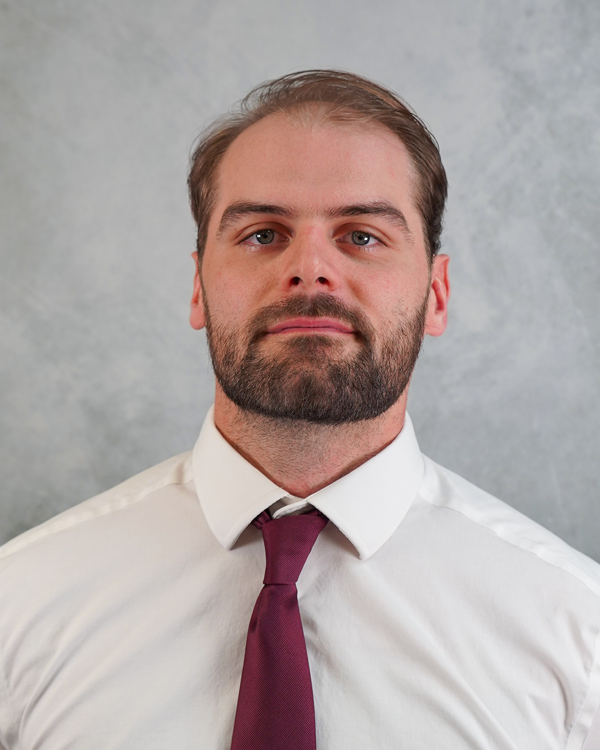 Zach Crosby poses for a headshot wearing a white shirt and red tie against a gray background