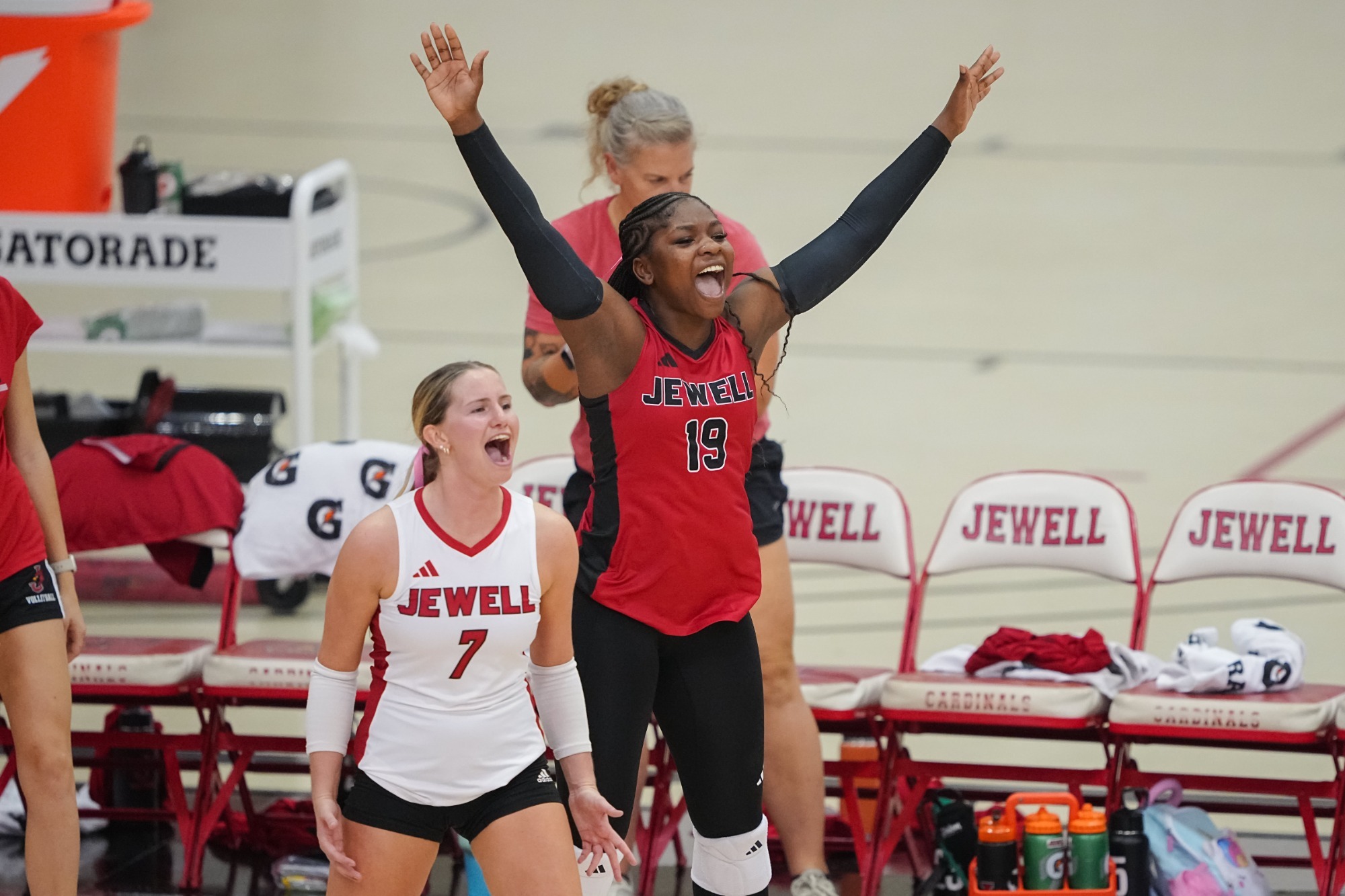 Alexis Frazier and Brianna Witthar celebrate a point in volleyball. Frazier is wearing a red volleyball jersey with her hands in the air while Withar has a white jersey on with an assistant coach in the background