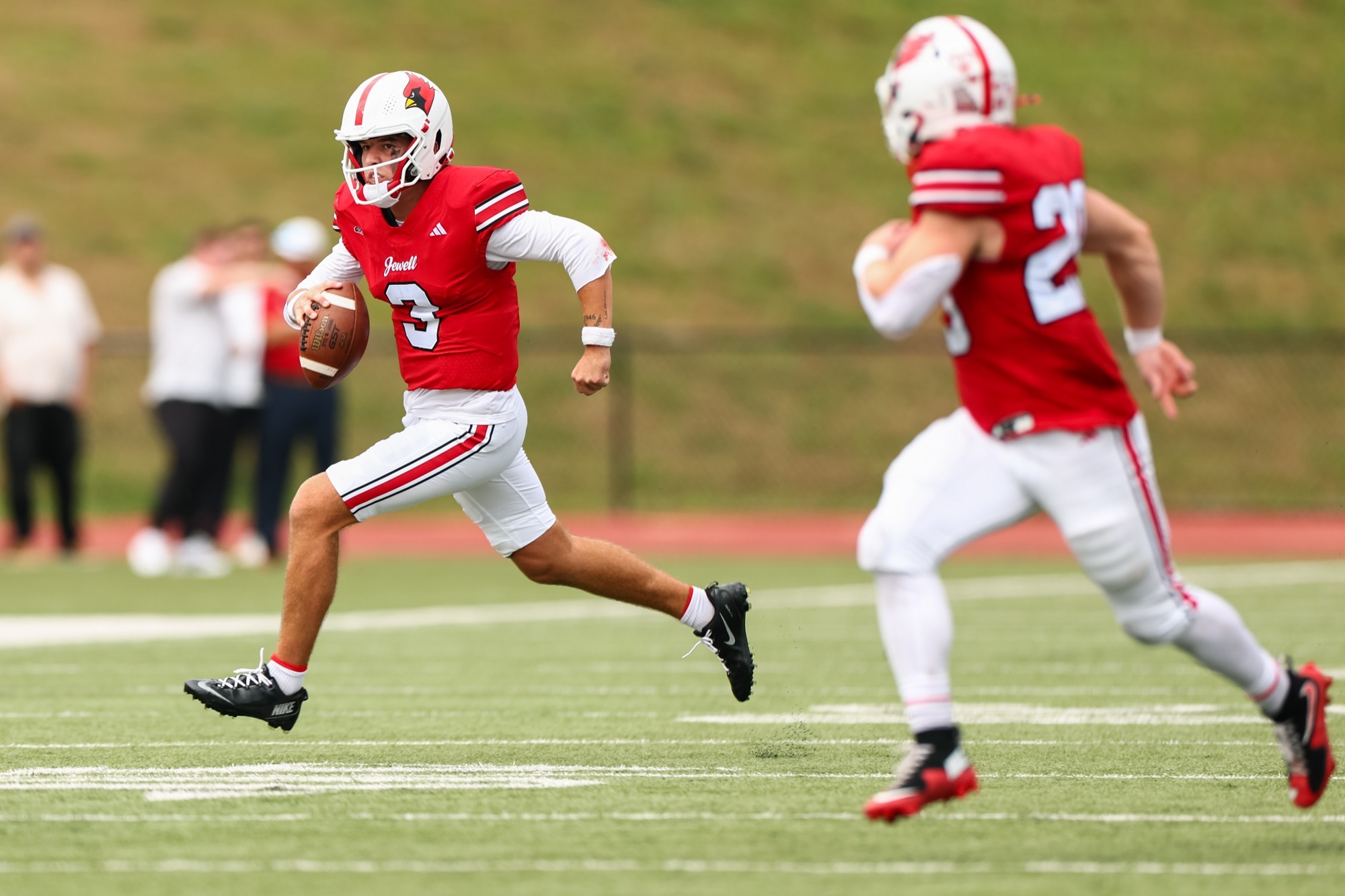 Charlie Loose, in a red football uniform with a white helmet, runs with the football in his right hand. Another Jewell player is blurred in the foreground