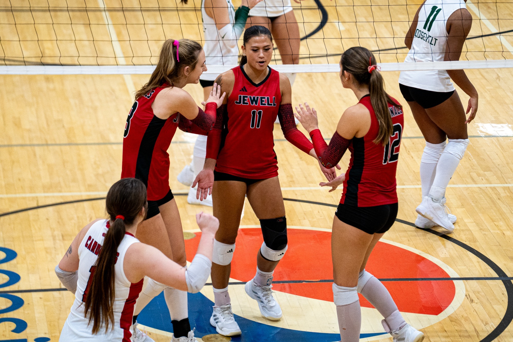 Silvia Fisher and Kali Adamec high five Lily Waters after scoring a point in volleyball. The players are standing near the net and wearing red jerseys with the net slightly blurred in the background.
