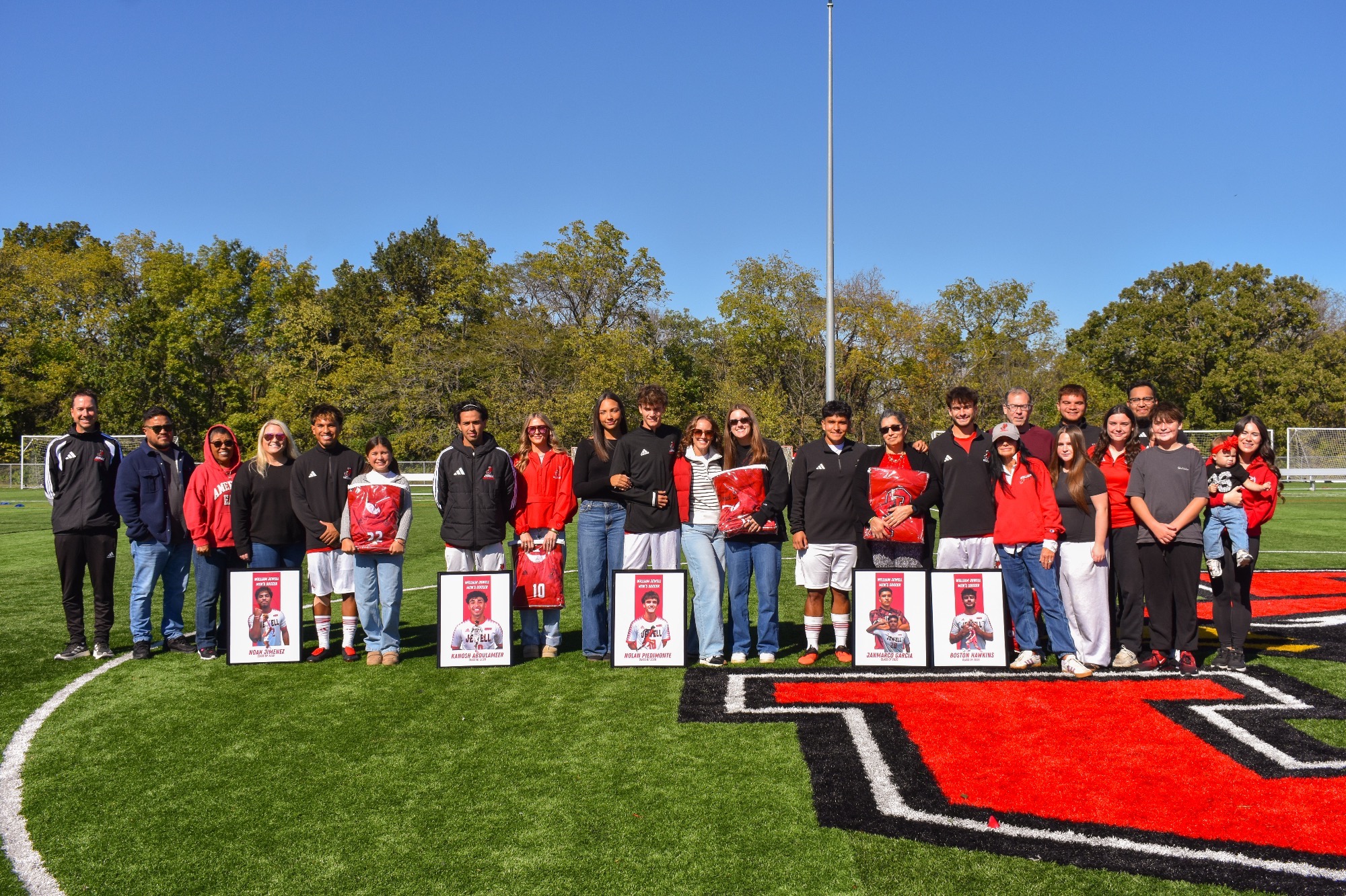 William Jewell men's soccer seniors pose with family, friends, and coaches on the field during Senior Day, holding framed player portraits and commemorative jerseys.