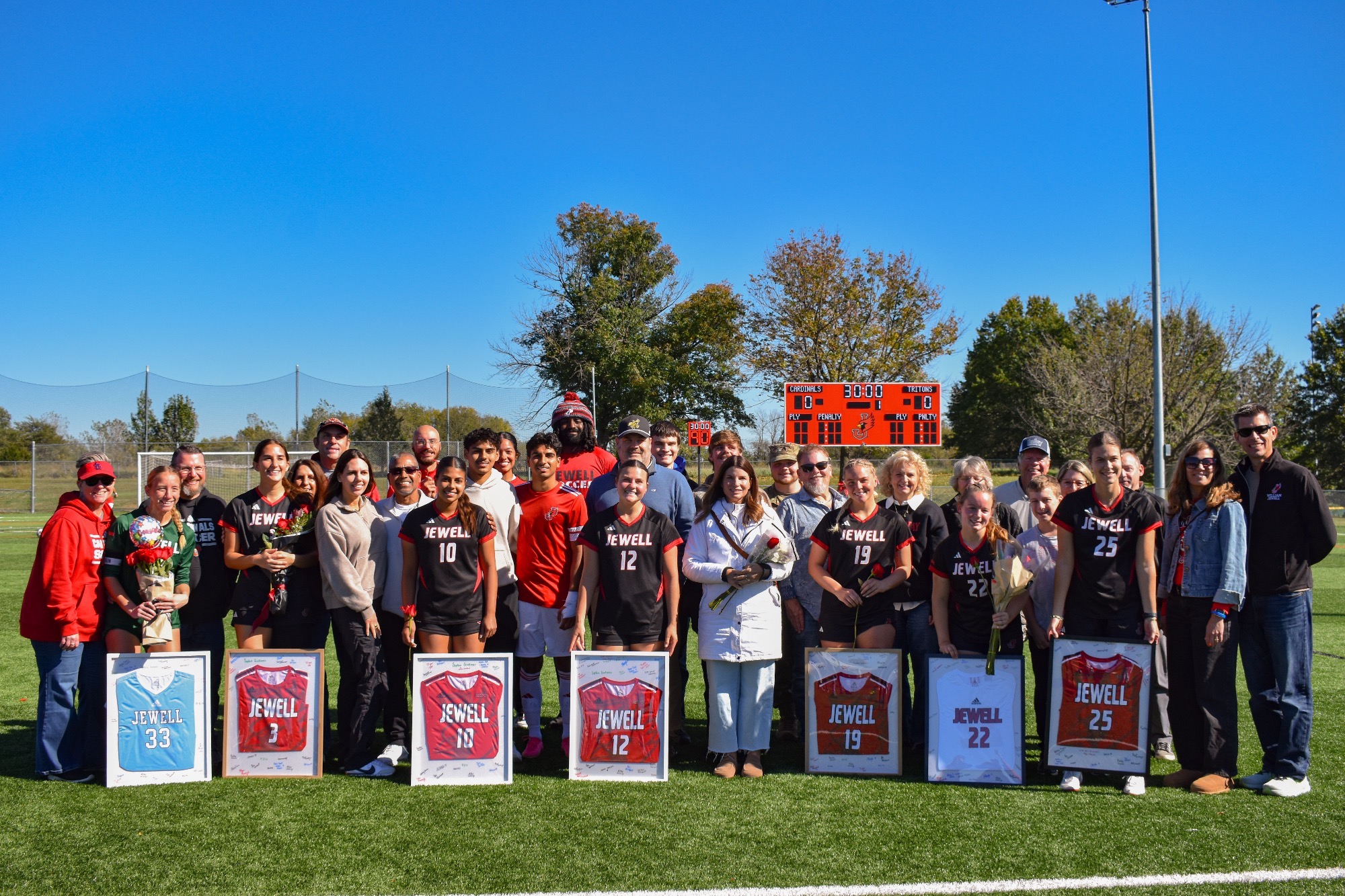 William Jewell women's soccer seniors pose with family, friends, and coaches on the field during Senior Day, holding flowers and framed commemorative jerseys in front of the scoreboard.