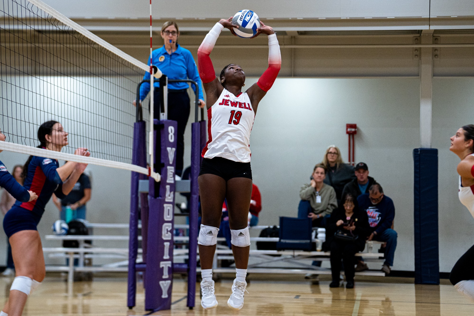 Alexis Frazier sets a volleyball in front of the net, wearing a white uniform. Small bleachers can been seen in the background.