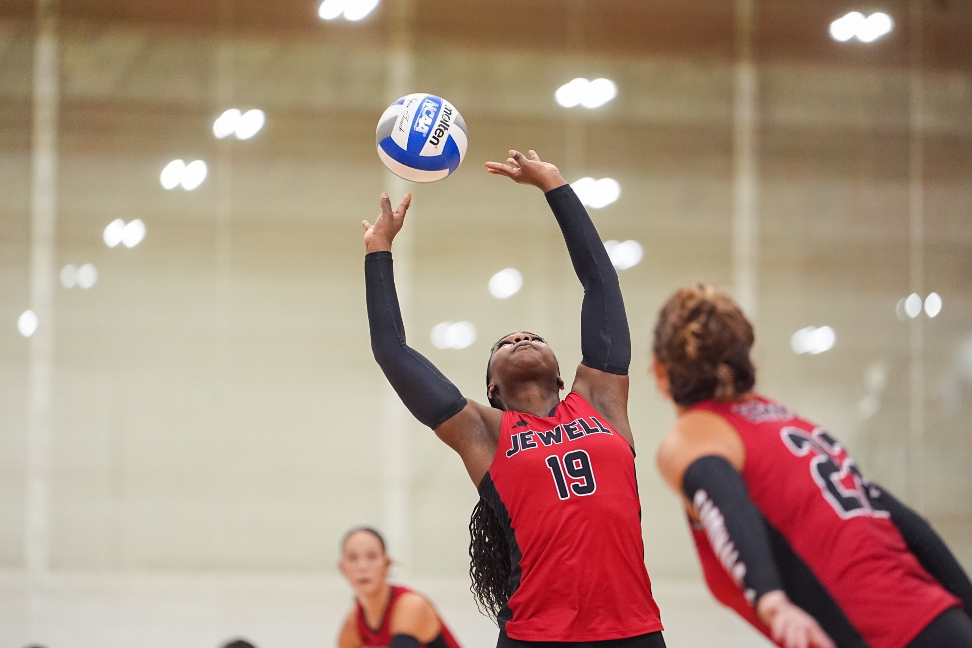 Alexis Frazier setting a volleyball behind her wearing a red uniform with one teammate blurred in foreground and one in the background