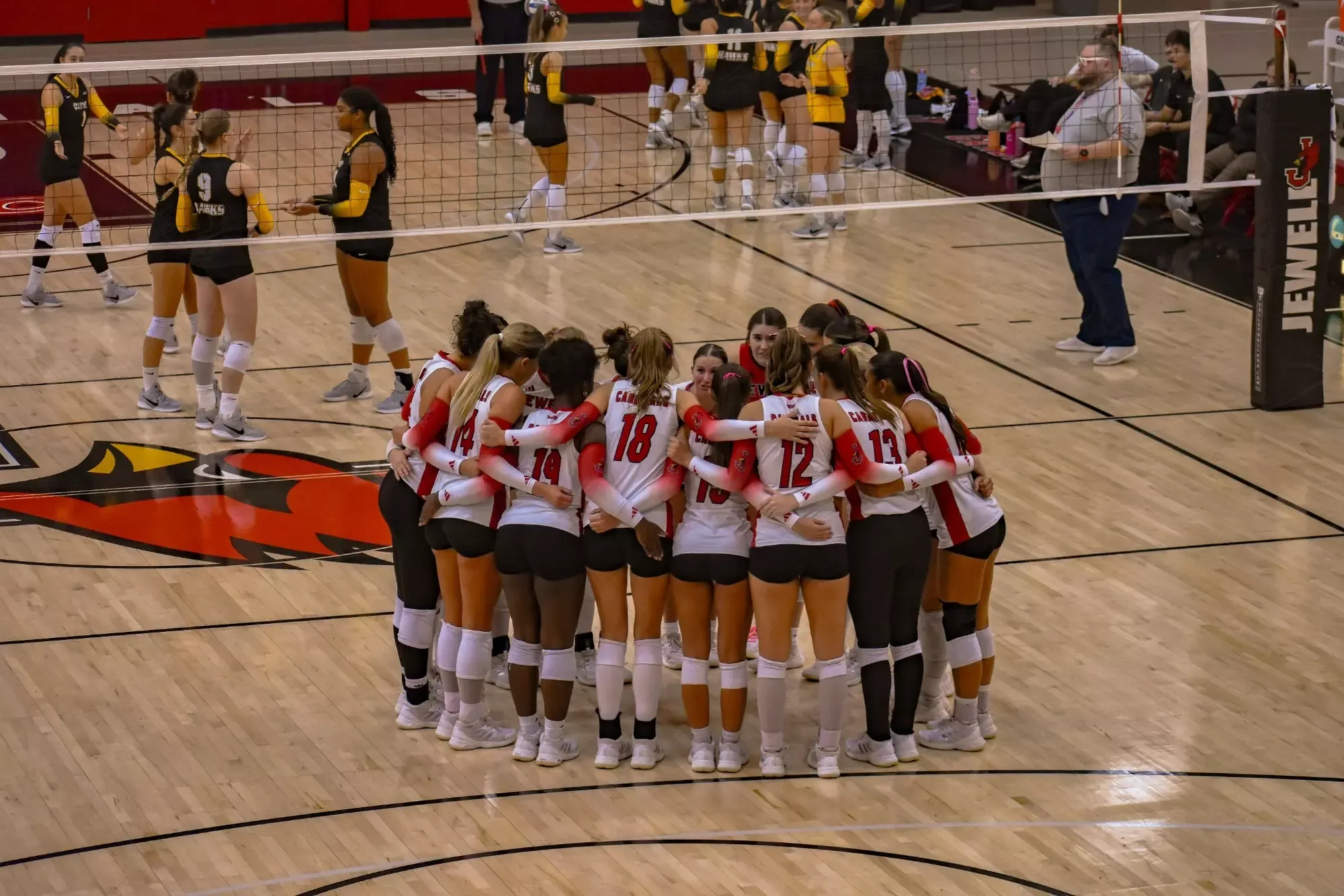 Members of the Jewell volleyball team stand in a huddle with their arms around each other prior to the start of a match. The opposing team can be seen in the background on the other side of the net.
