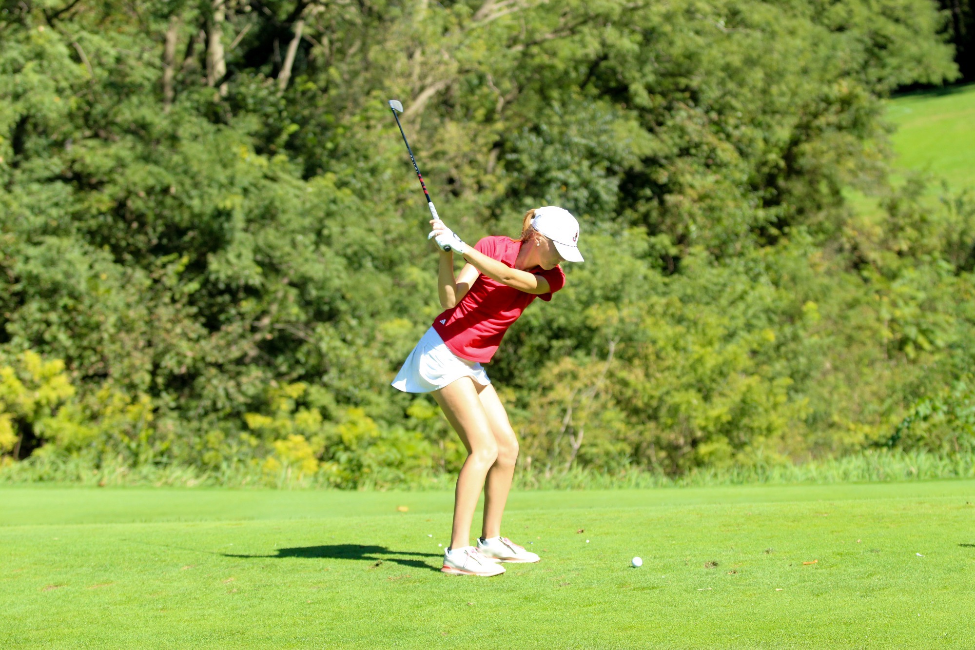 Scotlynn Blackington take a swing with a golf club, a green fairway behind her wearing a red shirt and white skirt.