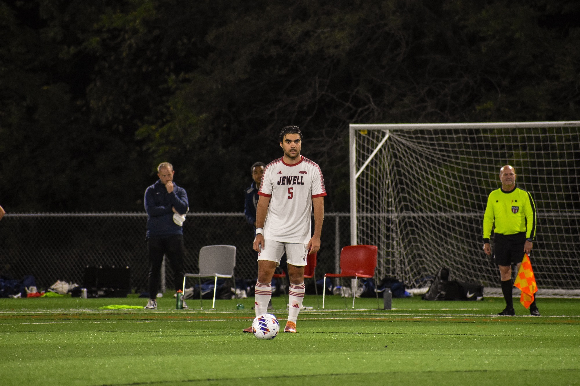 Giorgos Eleftheriou stands behind the soccer ball, looking where to pass to as two coaches and an official stand in the background slightly blurred.