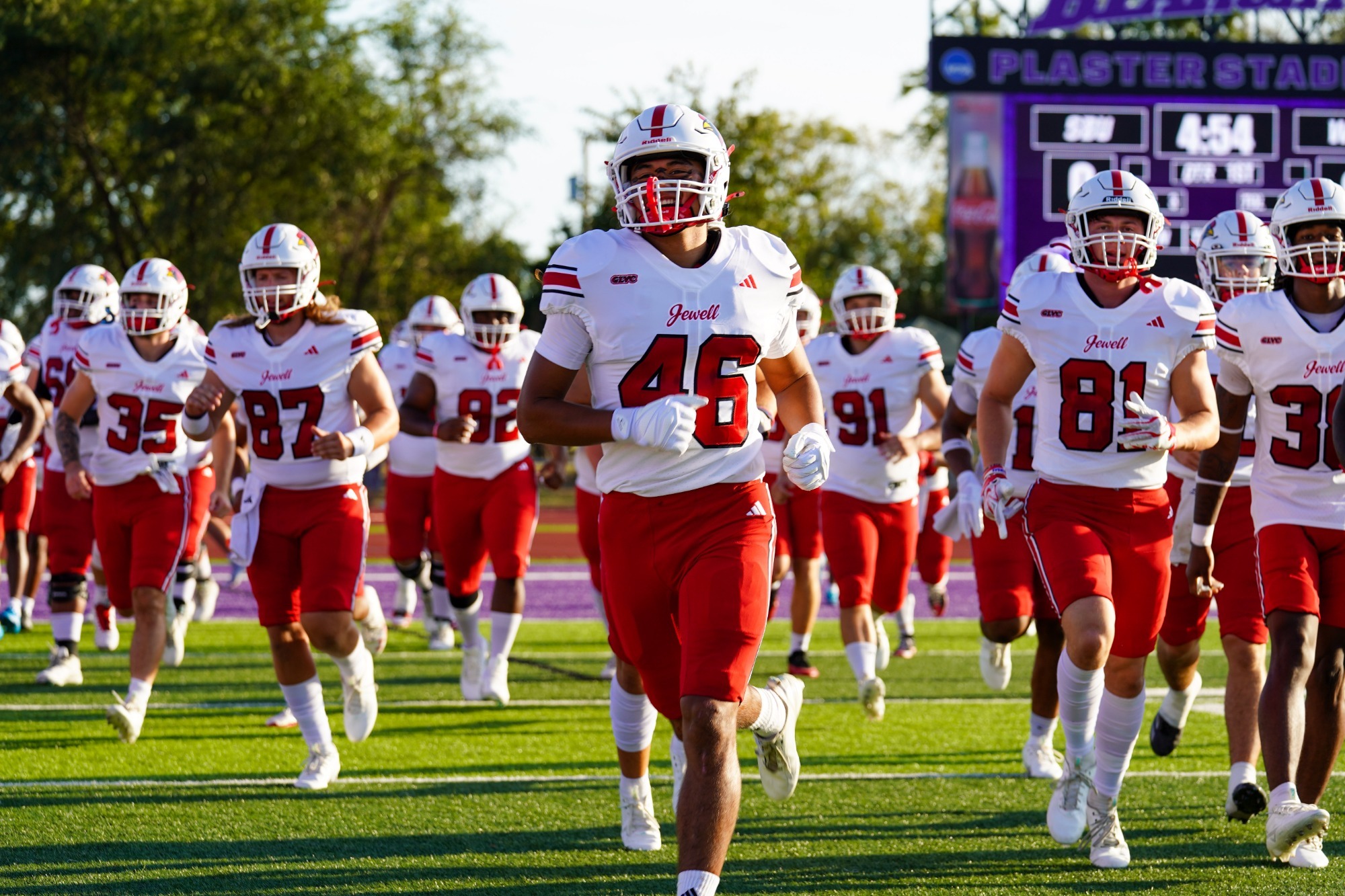 William Jewell football players in white jerseys and red pants run onto the field before a game, led by Sonny Proctor and Ben Podor
