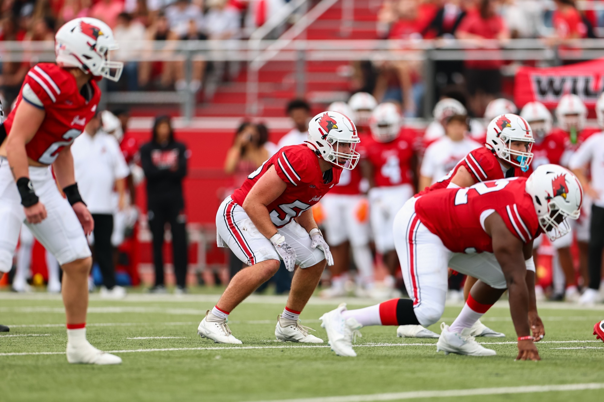 Jonny Williams and teammates in red jerseys, white pants and white helmets line up in formation on the field during a game, with teammates and spectators visible in the background.