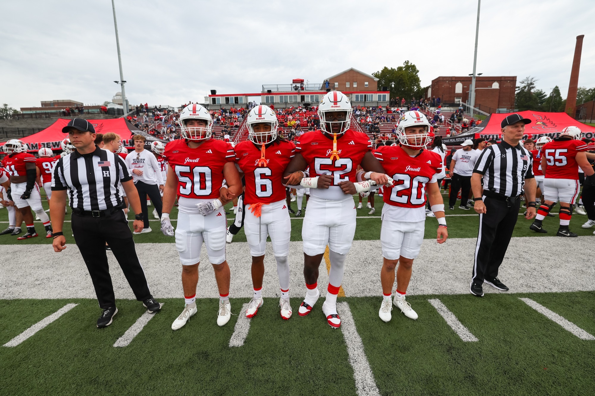 The four captains of the William Jewell football team, wearing red jerseys, white pants, and a white helmets stand with an official on each side ready to take the field for the coin toss. The bleachers and fans are visible in the background