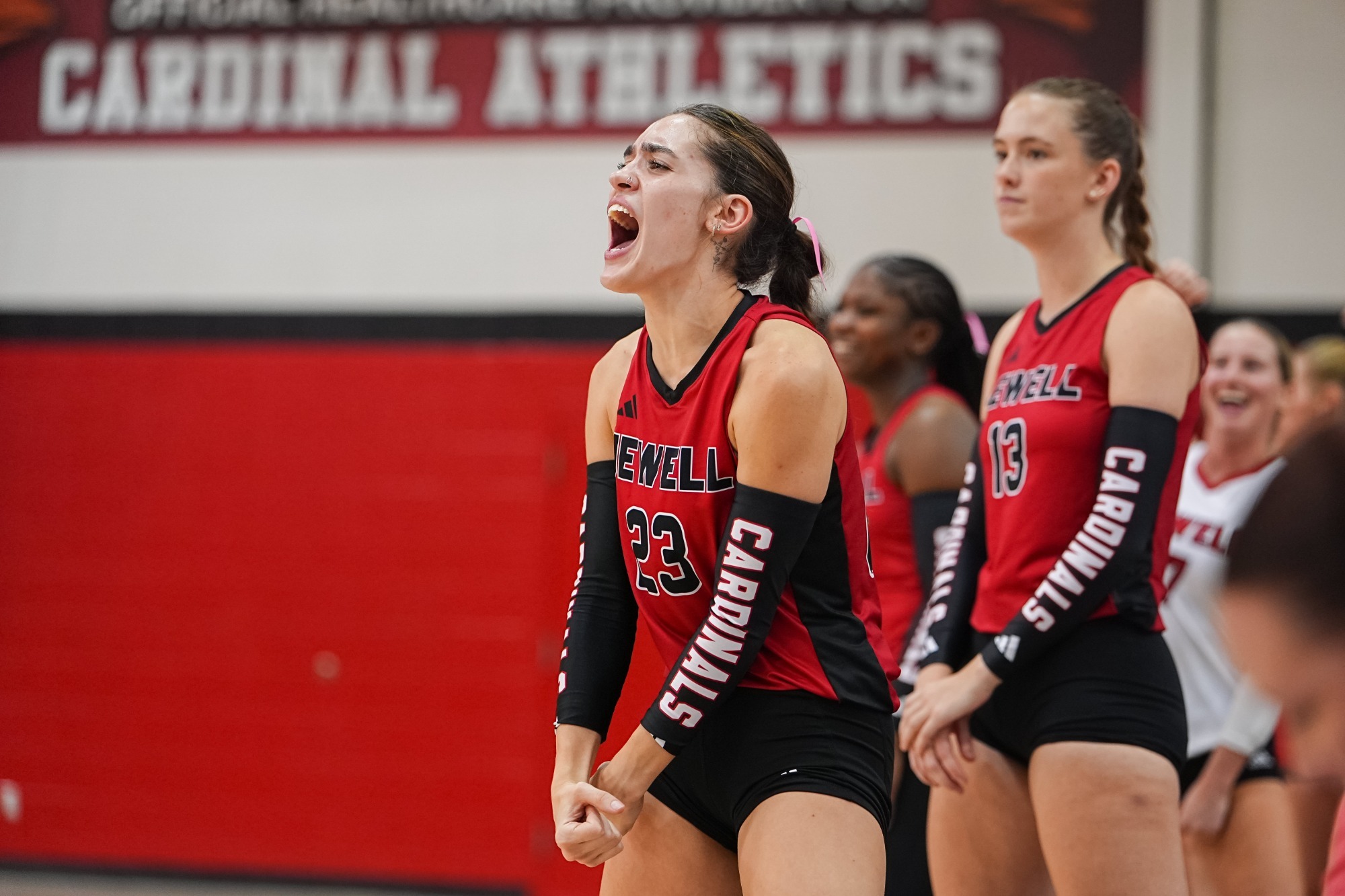 Olivia Starr celebrates a point, flexing her arms downward while yelling. She is wearing a red volleyball jersey with black sleeves. Teammates are standing behind her, slightly blurred.