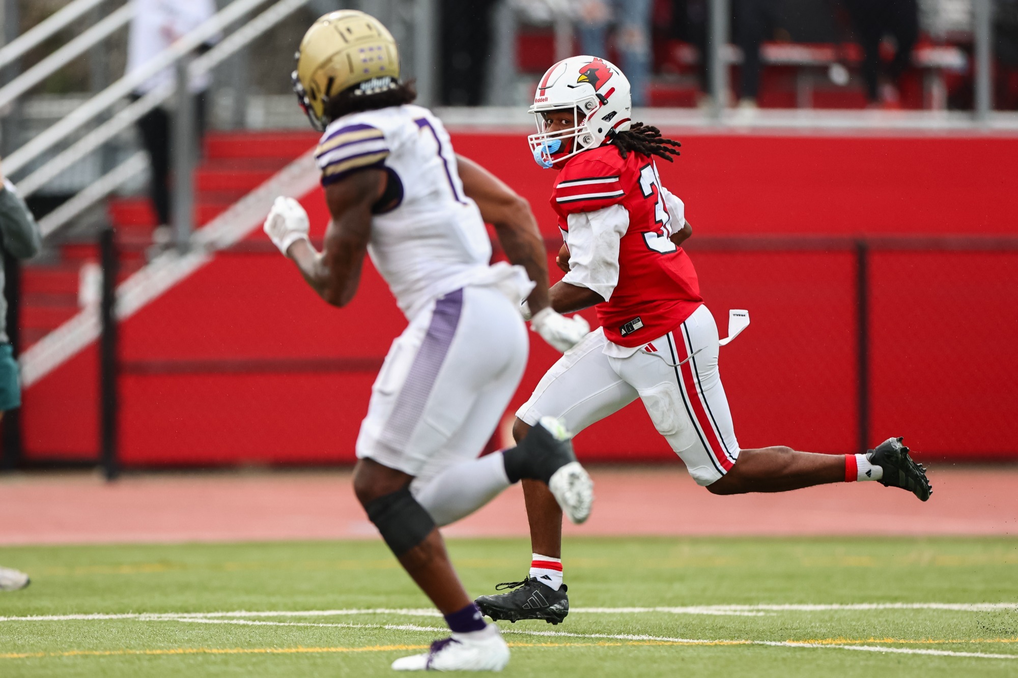 Kavious Record running with a football wearing red uniform, white pants, and white helmet with a red Cardinal head on the side. A McKendree player in a white uniform and gold helmet is slightly blurred in the foreground running.