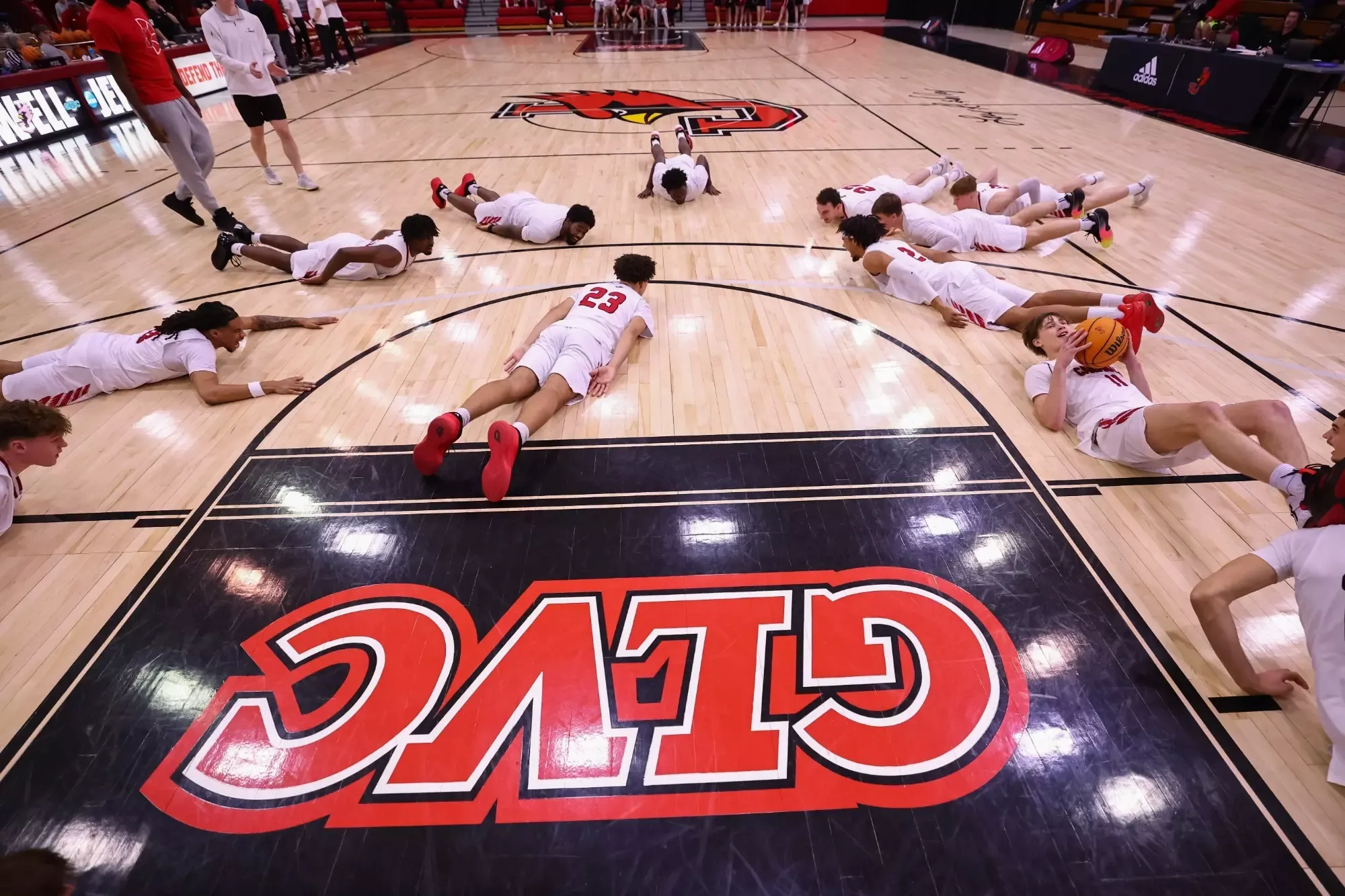 Jewell Cardinals men’s basketball players lie on the court in a circle during a pregame warmup, with one player holding a basketball. The team’s large Cardinals logo is visible at center court