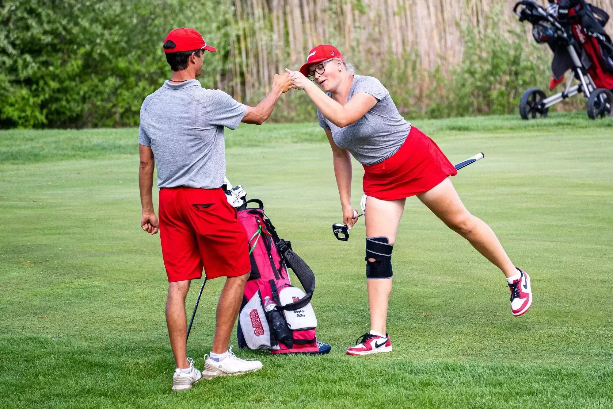 Brylie Ellis fistbumps her coach, Ian Barnes on a golf course. Both are wearing red shorts, gray shirts, and red hats