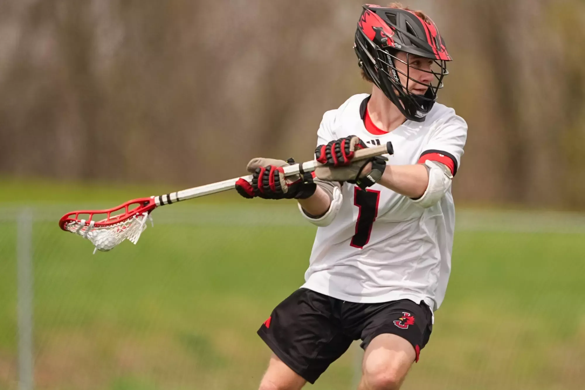 Nathaniel Leroux in a white jersey with the number 1 holding a lacrosse stick with a ball in the netting and looking to pass, wearing a black helmet and red gloves