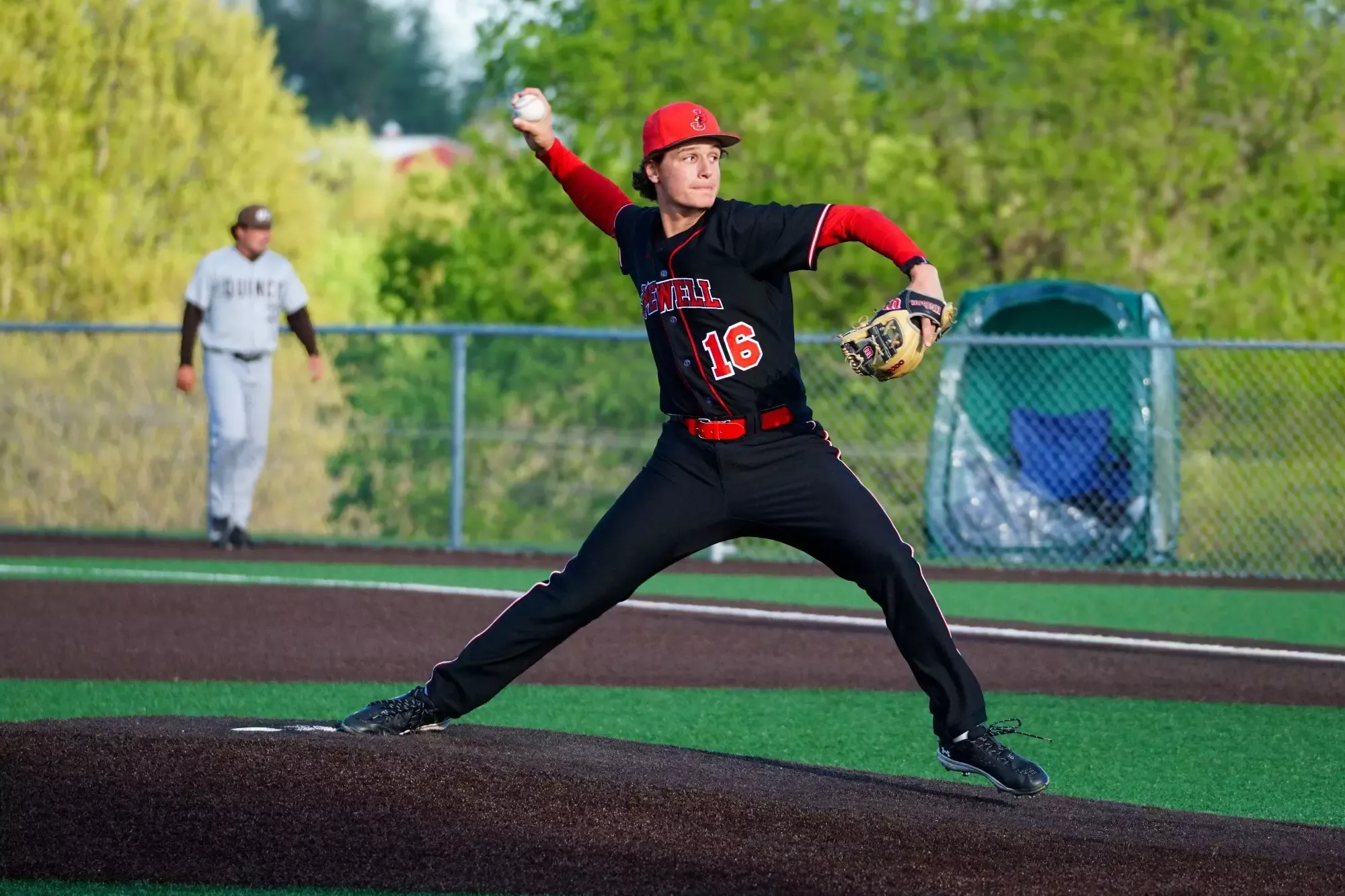Mike Wonderlich pitches a baseball while wearing a black uniform, red sleeves, and red hat. A Quincy player in a gray uniform is blurred walking in the background in front of trees and fencing