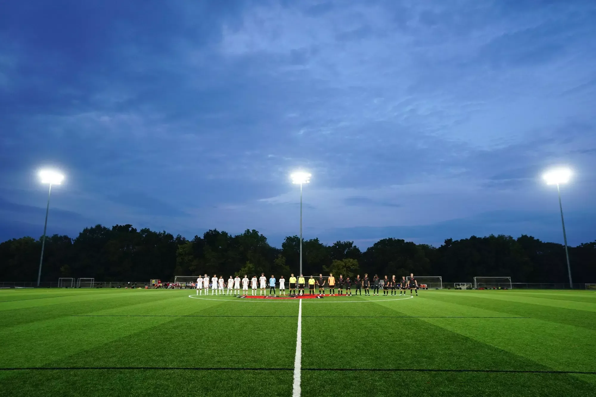 William Jewell men's soccer players and opponents line up at midfield before a night match under the stadium lights