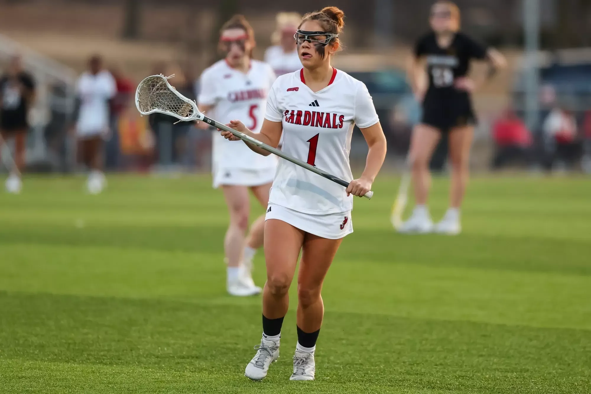 Toryn Podschwidt runs while holding a lacrosse stick and wearing an all white uniform and goggles. Other players are blurred in the background