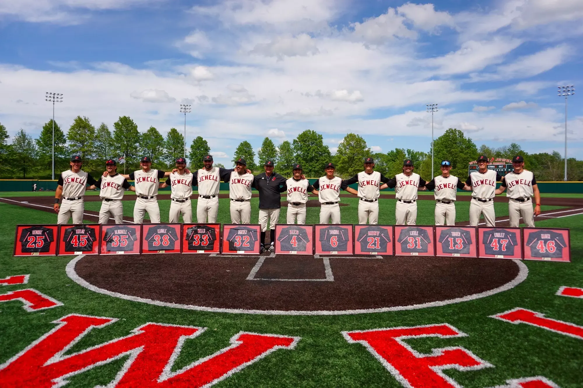 13 seniors are standing in a line with their arms around each other with head coach Mike Stockton standing in the middle. All players are wearing a cream uniform with Jewell across the chest and black hats with framed black jerseys on the ground in front of them. A clear blue sky is visible with trees and light poles in the background