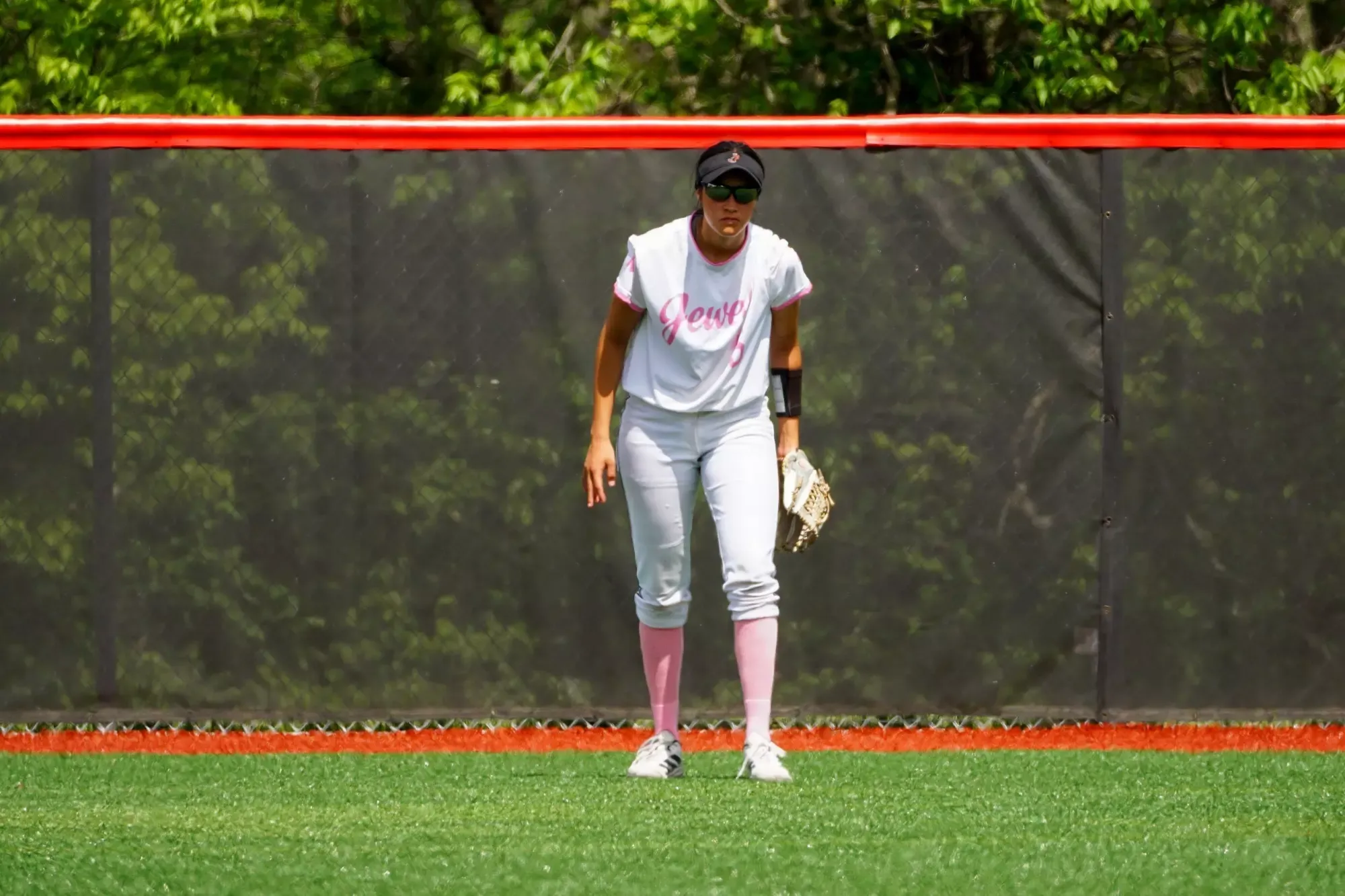 Mikayla Chairez, wearing an all white uniform with pink trim, pink socks, and a black visor and sunglasses stands in center field in front of a black fence