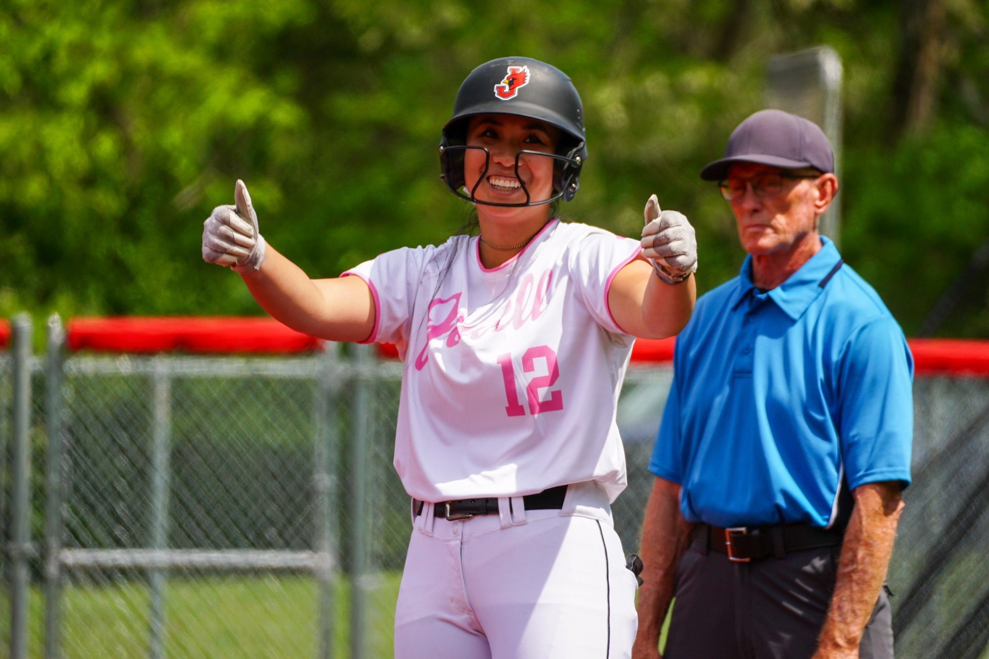 Softball player wearing white uniform, pink trim and black helmet giving thumbs up