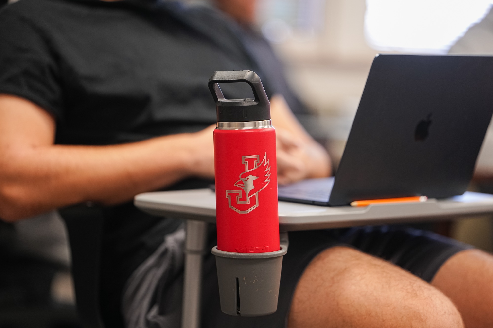 A red YETI water bottle featuring the William Jewell Cardinals logo sits in a cup holder attached to a classroom desk. In the blurred background, a student wearing a black shirt works on an open laptop during class.