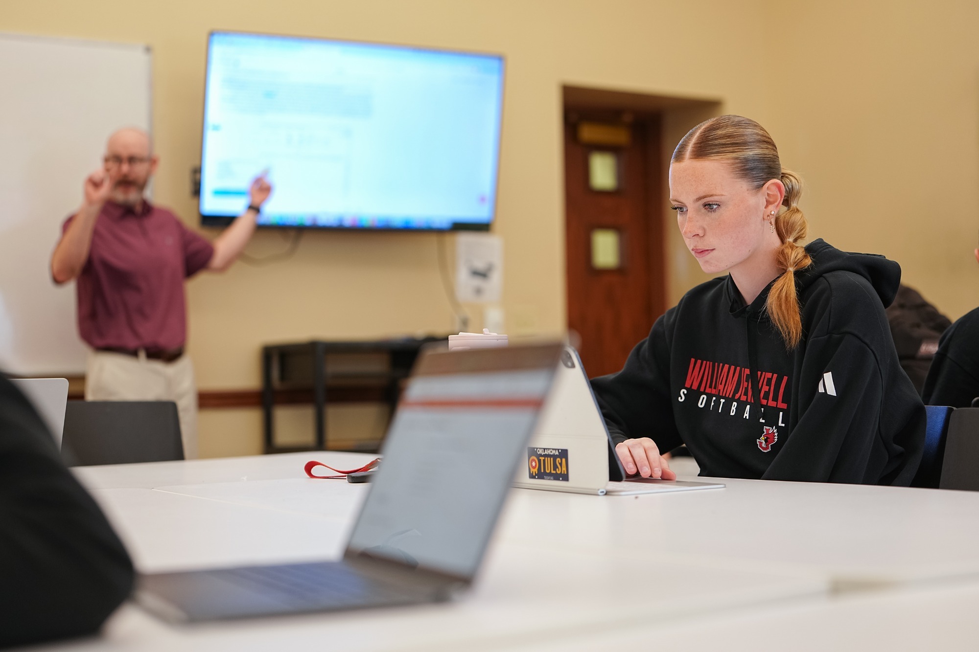 Student wearing a William Jewell Softball sweatshirt works on a tablet during a classroom session. In the background, an instructor gestures toward a presentation displayed on a screen while teaching. Other students and laptops are visible in the foreground, creating a collaborative learning environment