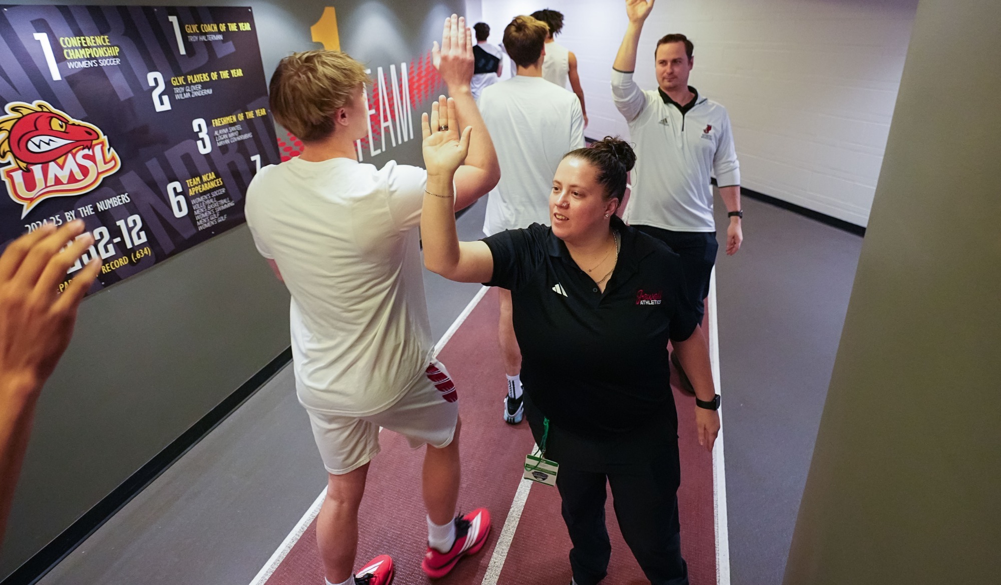William Jewell men’s basketball players walk through a hallway exchanging high-fives with athletics staff and coaches before a game. Team members wear white warm-up attire, while staff members stand along the walkway offering encouragement. Championship and program achievement graphics are displayed on the wall beside them