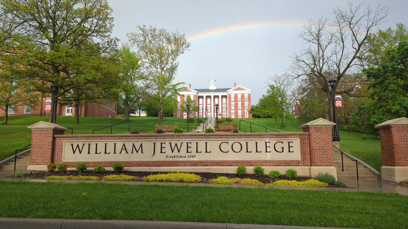 View of the William Jewell College entrance sign on a landscaped campus lawn, with the historic Gano Chapel centered on a hill in the background. A rainbow stretches across the sky above the campus following a rain shower, framed by trees and green spring foliage