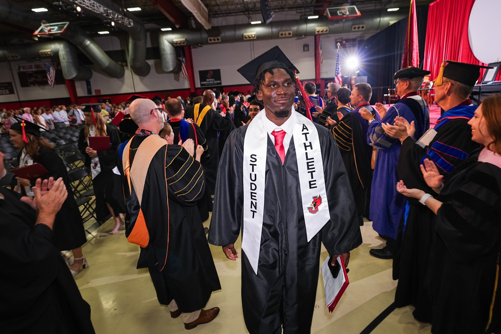 Student-athlete wearing a graduation cap and gown walks through an honor guard of faculty and staff during William Jewell College commencement. The graduate wears a white ‘Student Athlete’ stole and carries a diploma cover while faculty members in academic regalia applaud on both sides. The ceremony takes place inside a packed campus arena
